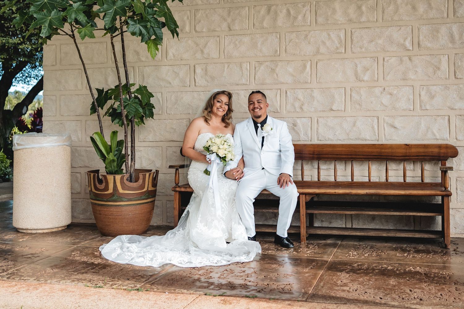 Newlyweds pose on wooden bench against stone wall in formal wedding attire.