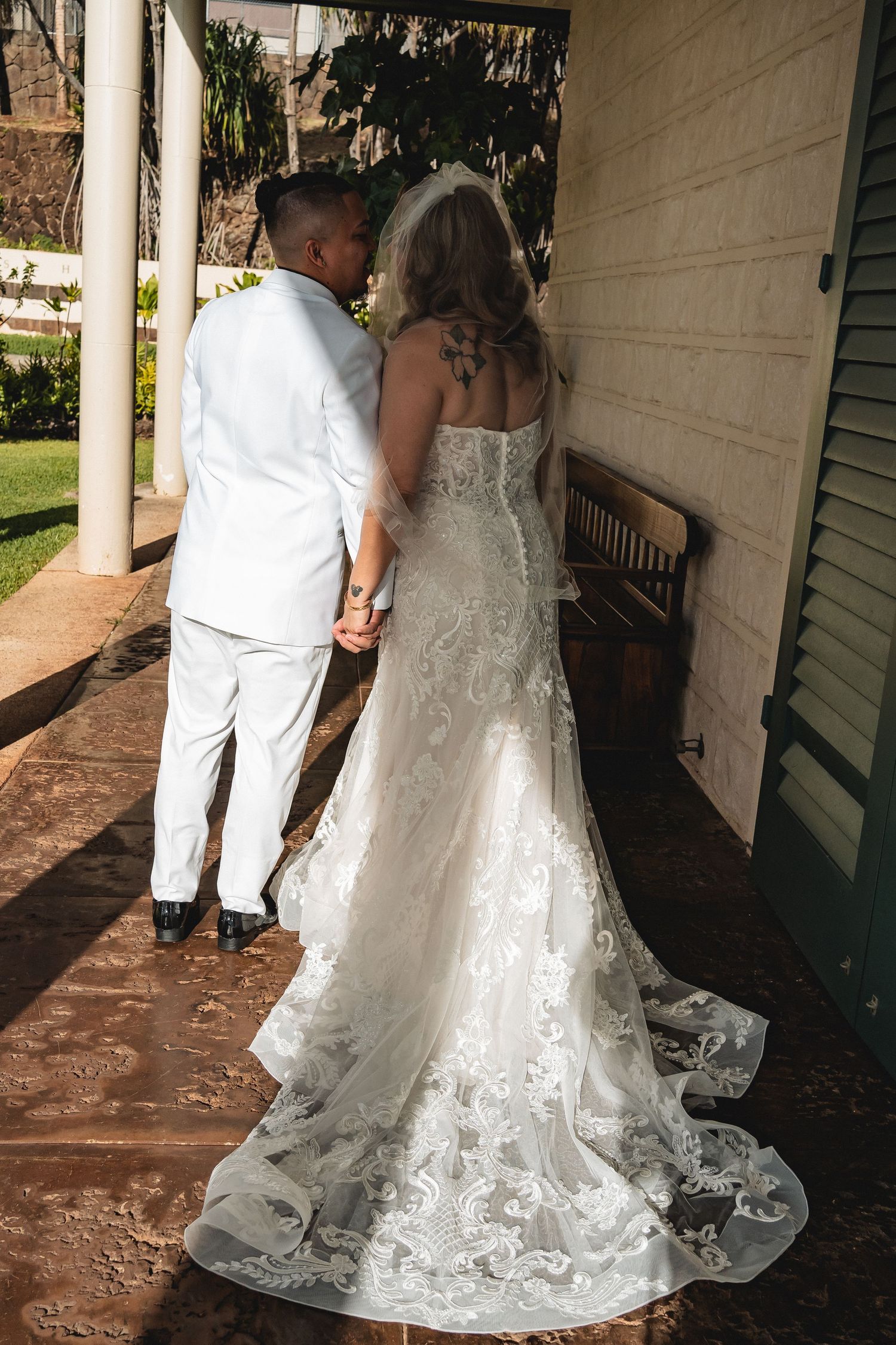 A wedding couple in white attire walking along a covered porch with intricate lace dress details.
