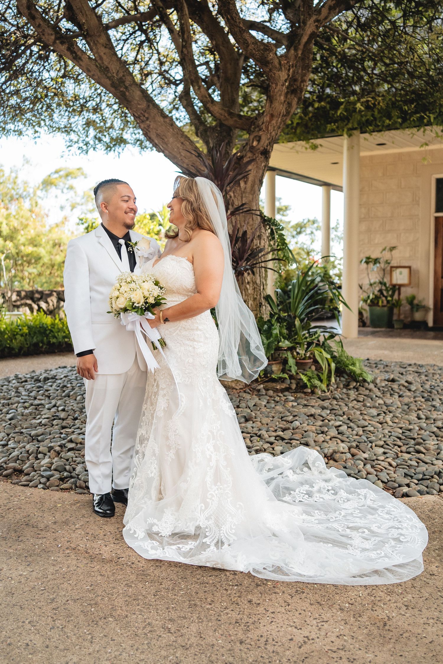 A wedding couple poses under a large tree with the bride wearing a flowing lace gown with long train.