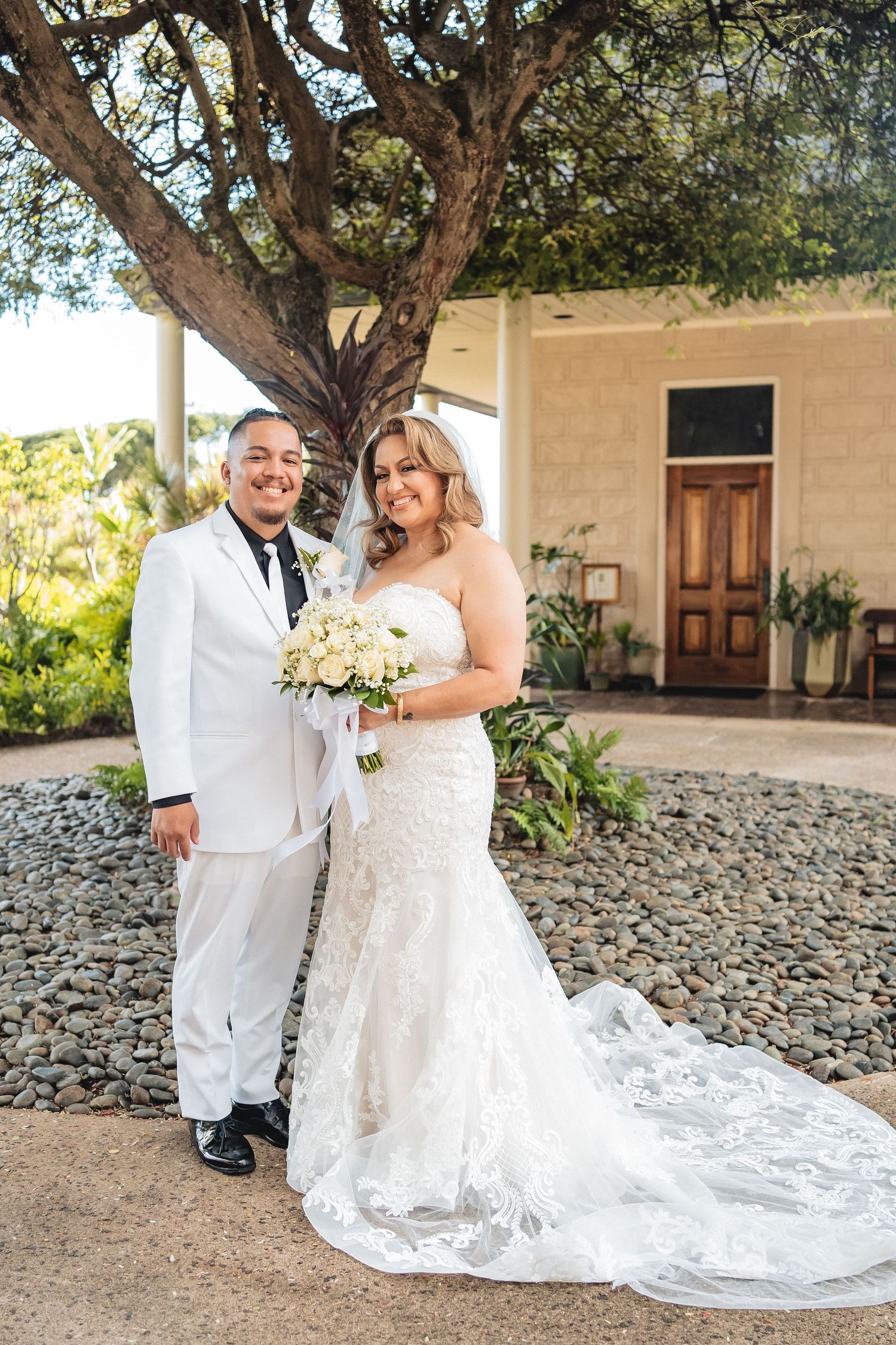 Wedding couple poses under large tree with stone building and landscaping in background.