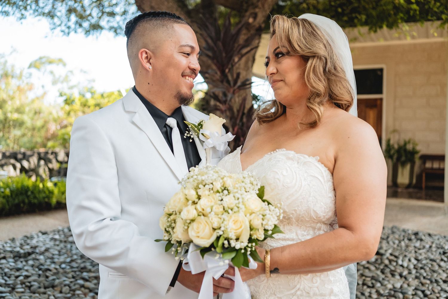 Newlyweds share a joyful moment outdoors while holding a bouquet of white roses.