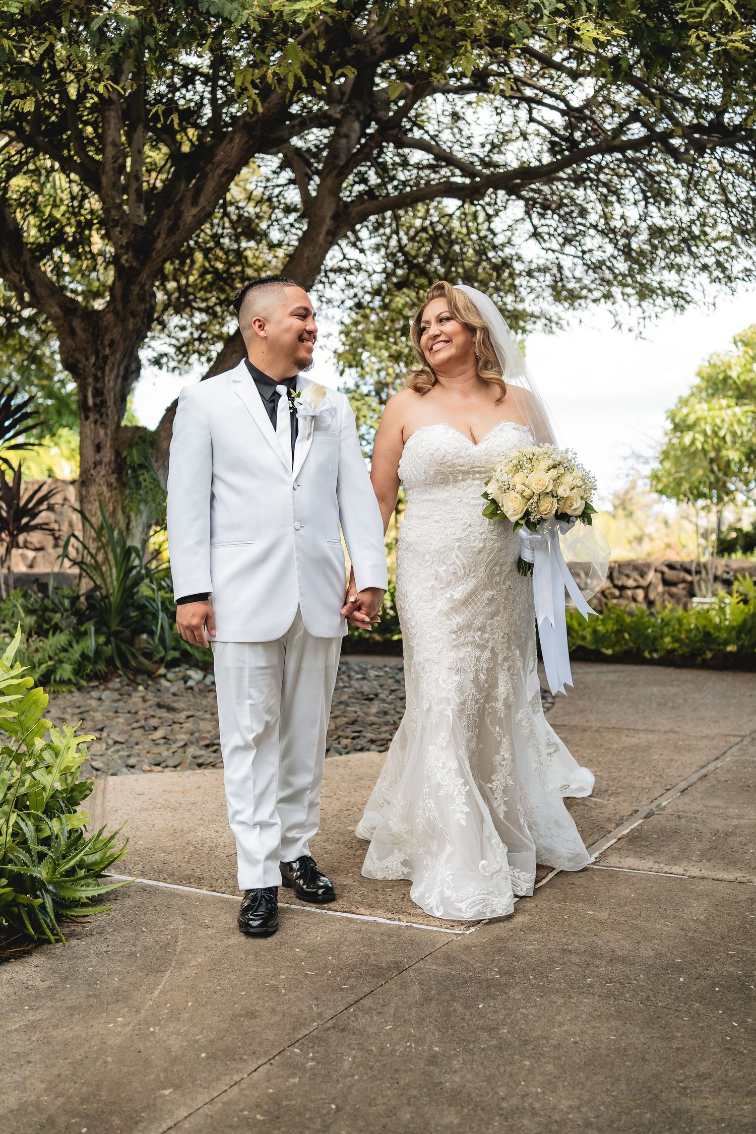 Wedding couple walks hand in hand along a garden path wearing white formal attire.