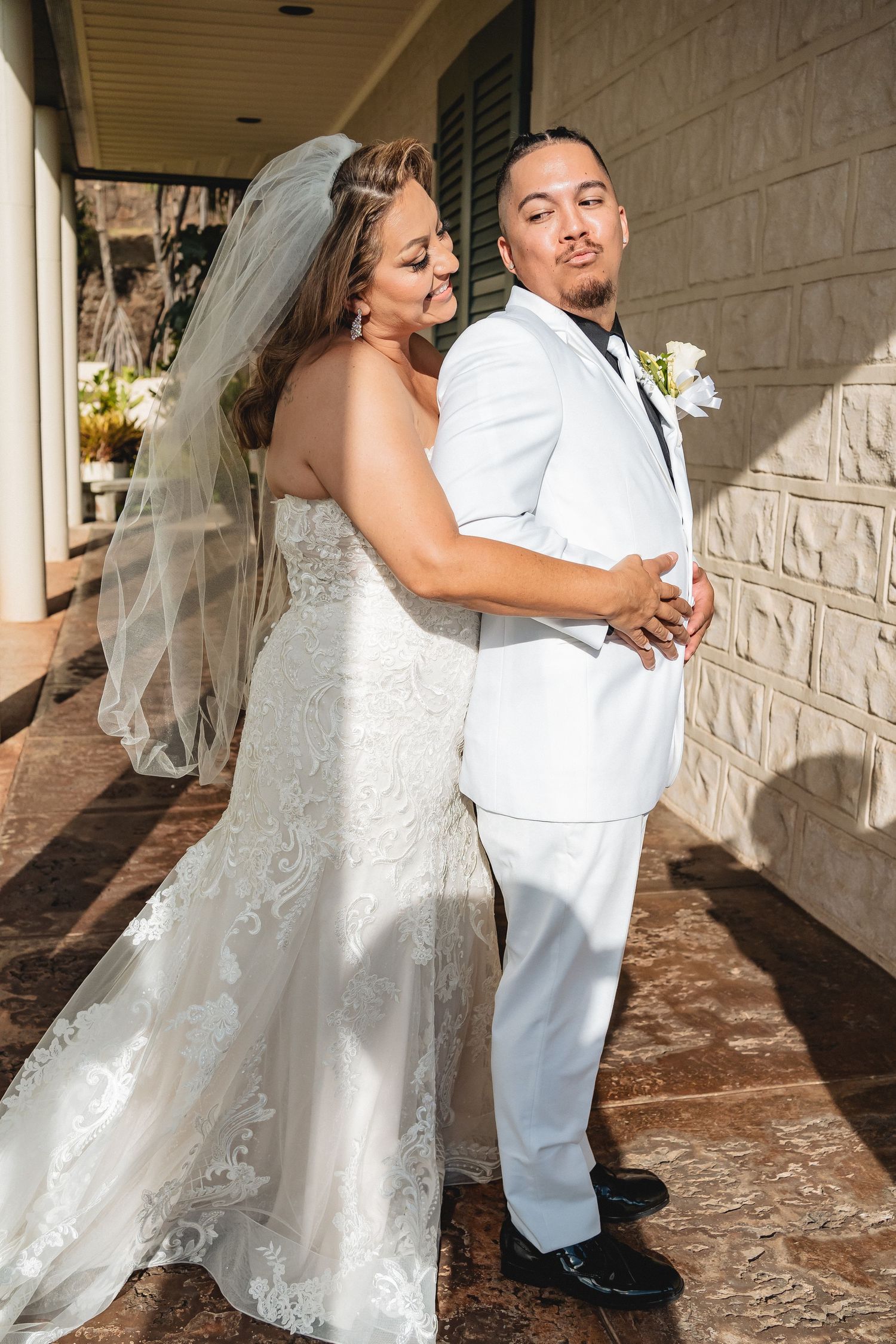 Playful wedding couple poses against white stone wall in elegant attire.