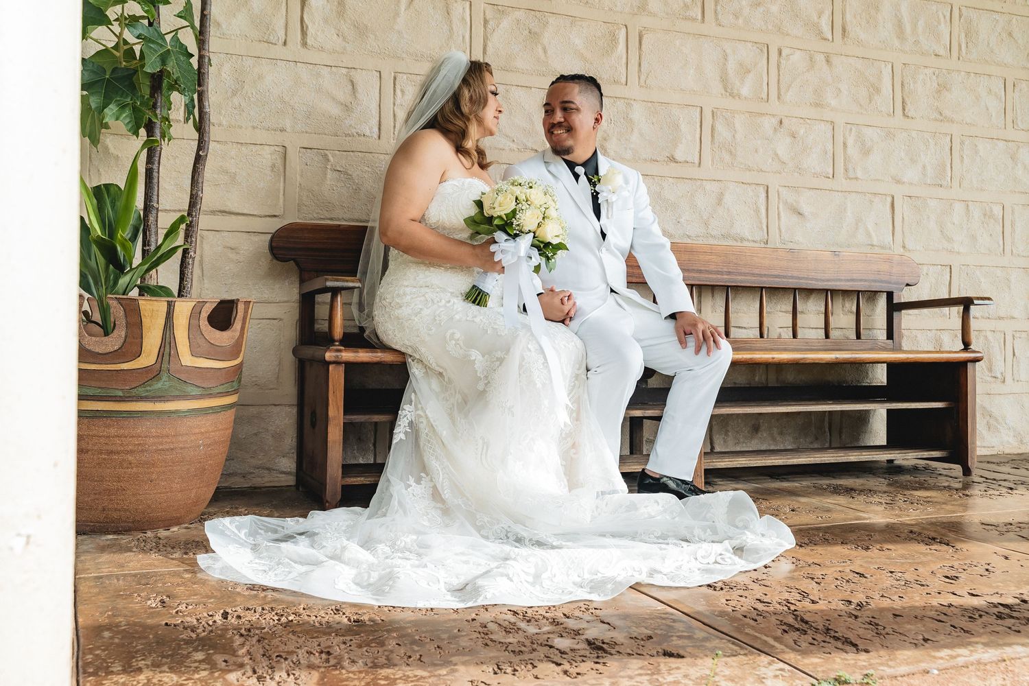 A wedding couple shares a moment on a bench with the bride holding a white floral bouquet.