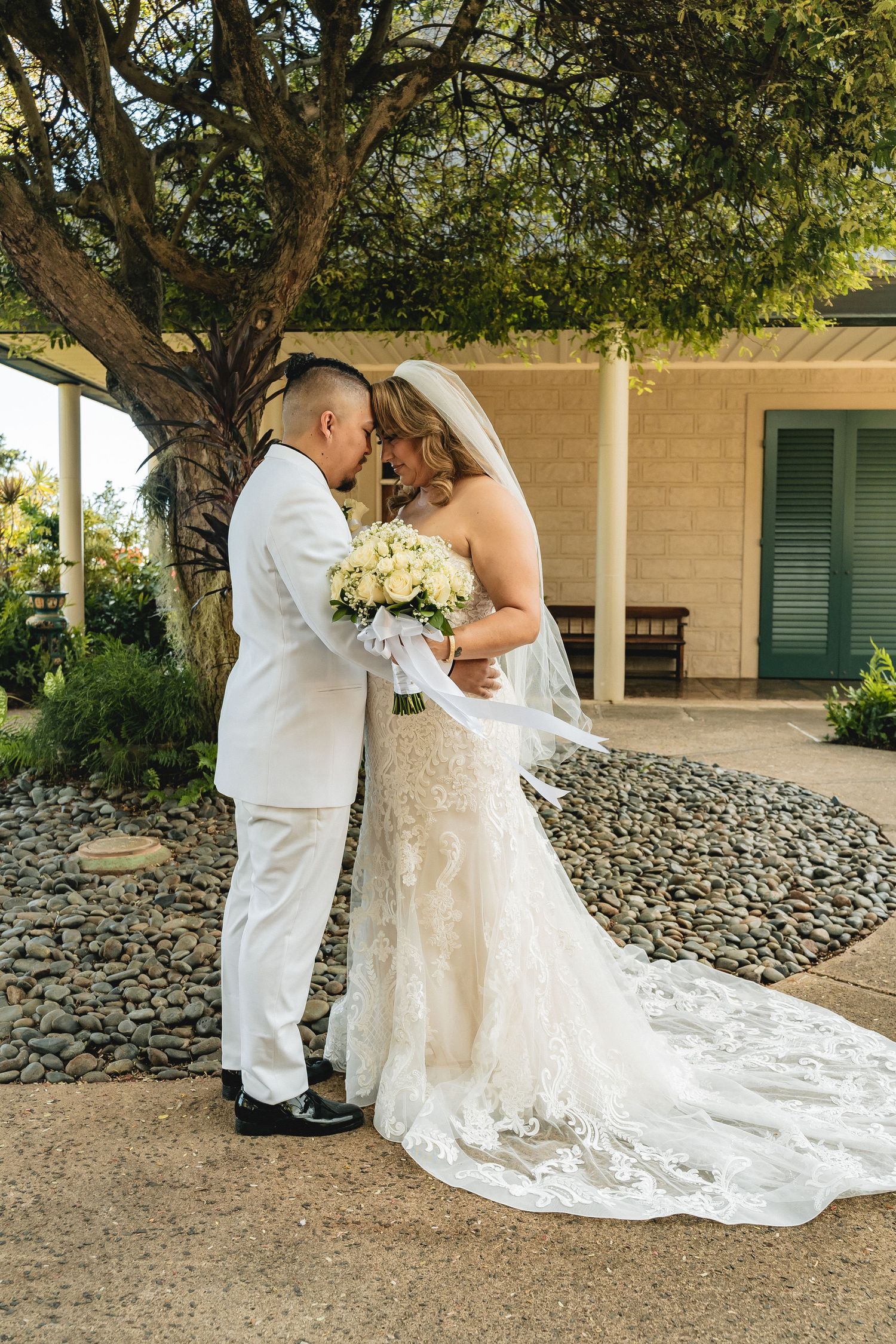Wedding couple embraces under large tree with bride's lace dress train flowing behind.