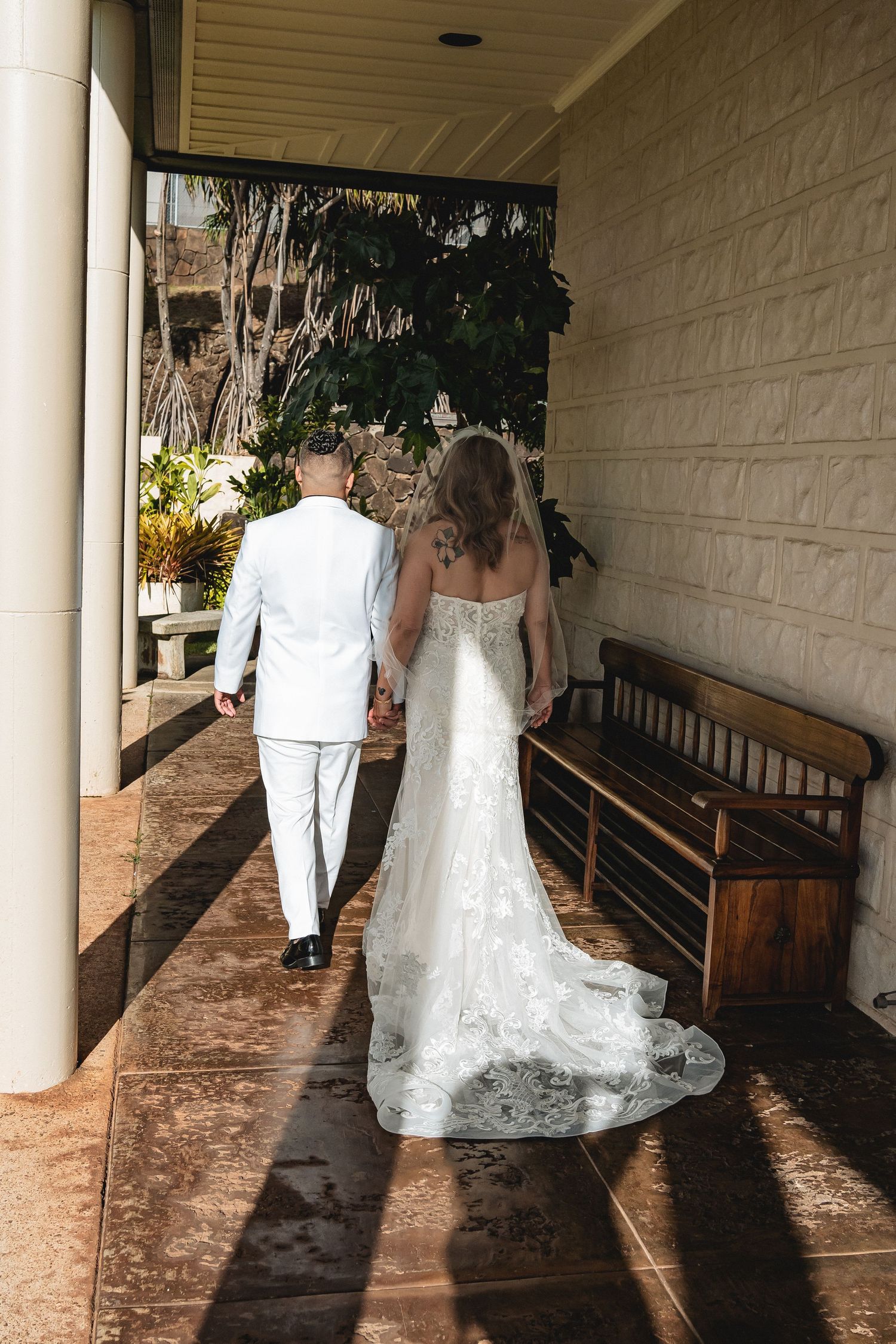 A newlywed couple walks down a covered walkway with the bride's lace train flowing behind.