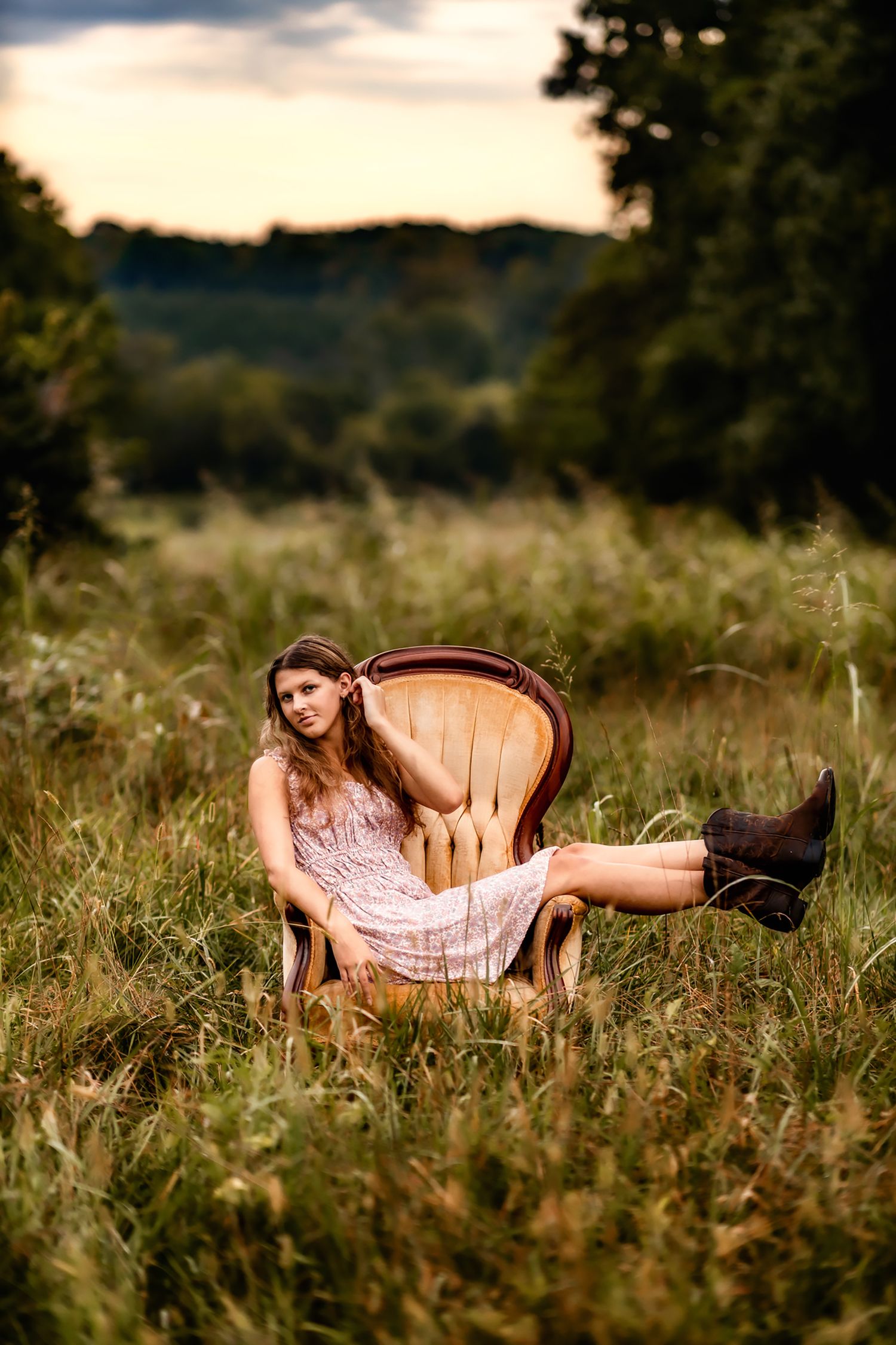 Woman in pink dress lounging in vintage chair placed in rustic outdoor field setting.