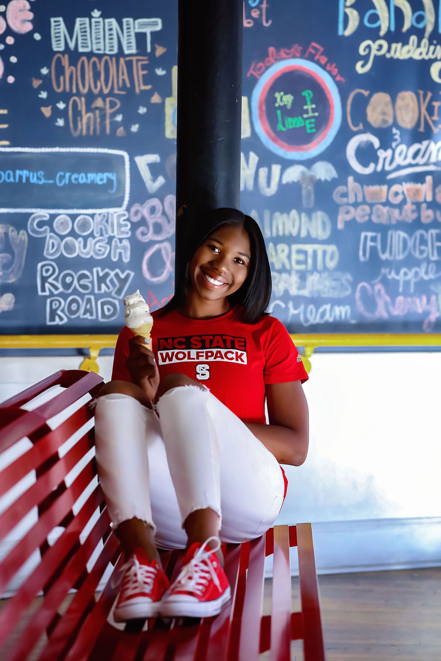 Student in red NC State Wolfpack shirt relaxing on red bench near chalkboard menu.