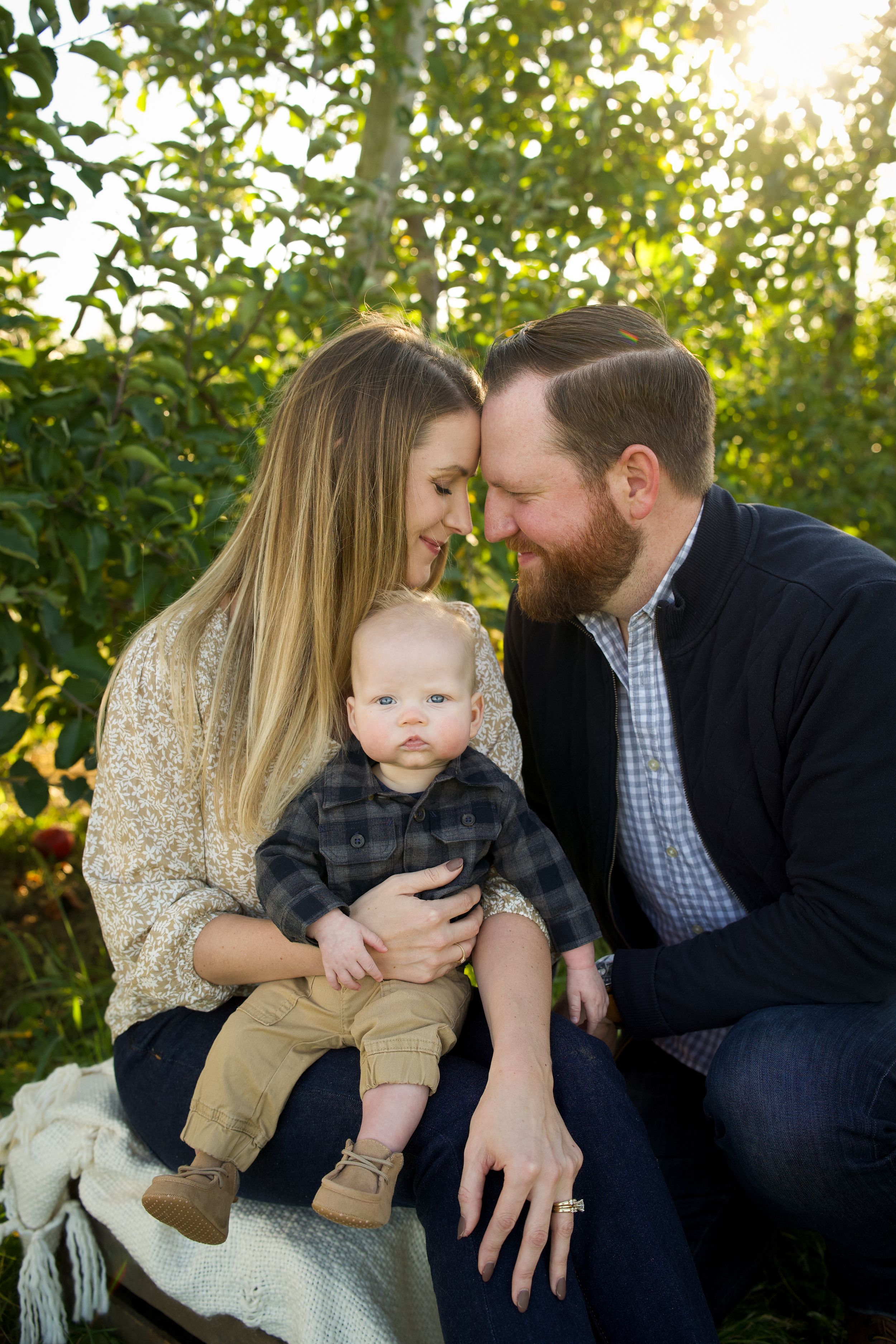 A tender family moment captured outdoors with sunlight filtering through trees in the background.