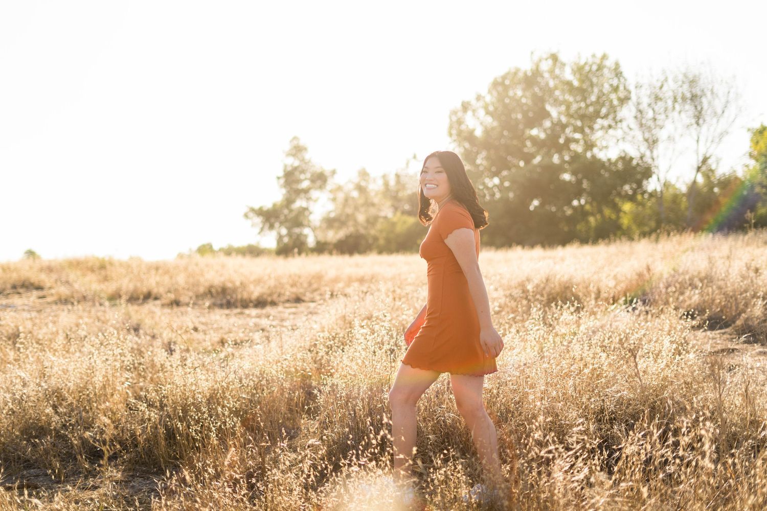 Silhouette of a high school senior in an orange dress walking through a golden sunlit field during sunset.