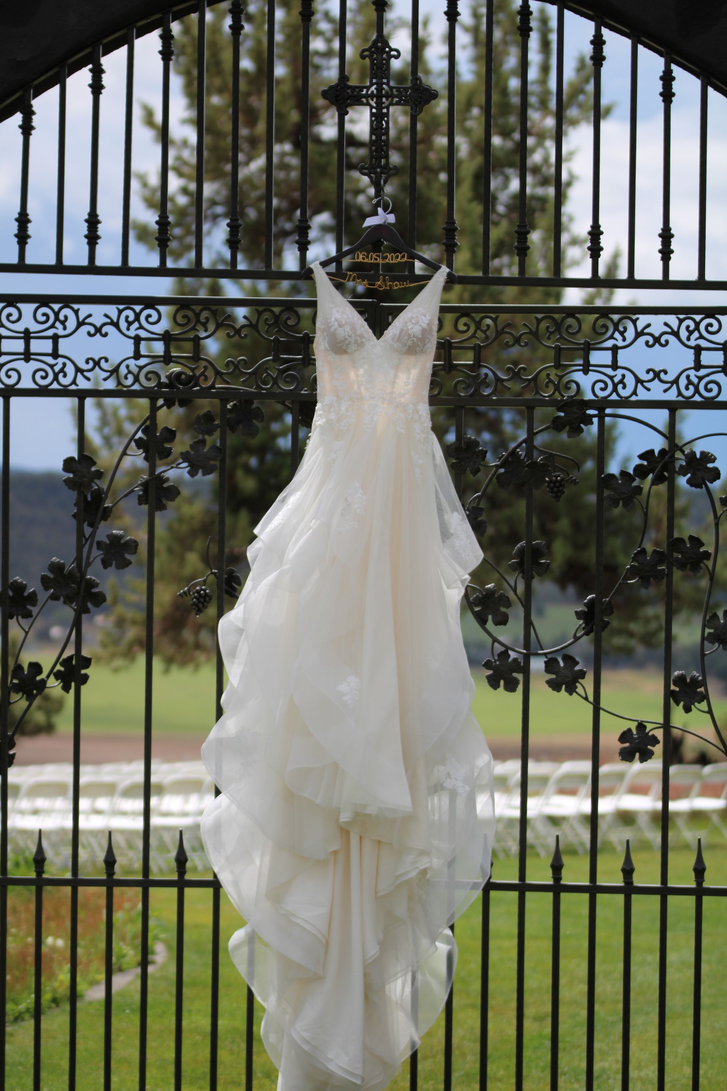 White wedding dress hanging on ornate iron gate with garden view in background.