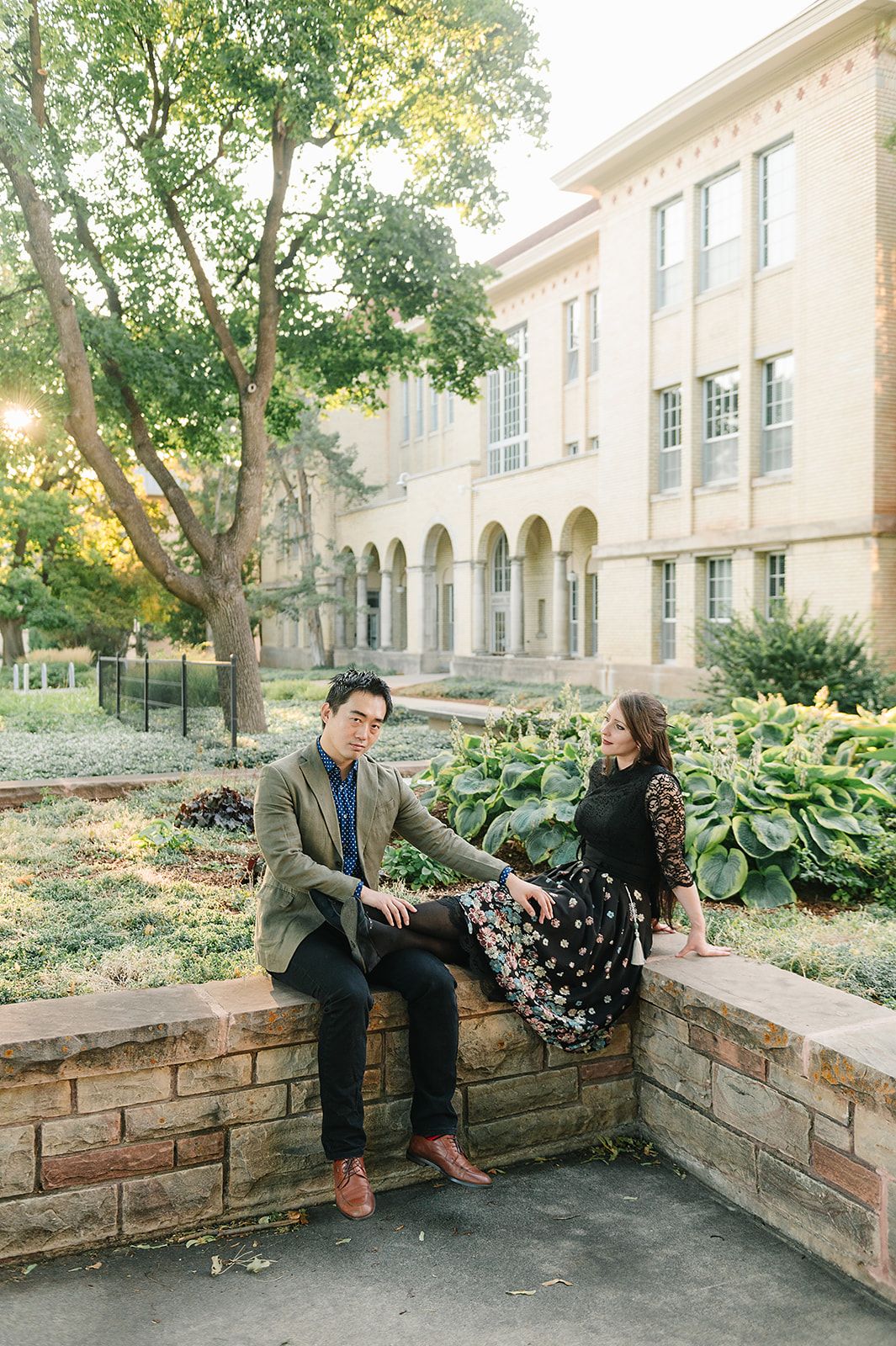 A couple sits on a stone wall outside a classic academic building with arched windows and lush gardens.