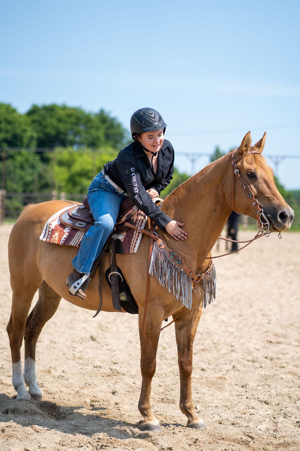 Western rider on a dun horse with fringed saddle in competition arena.