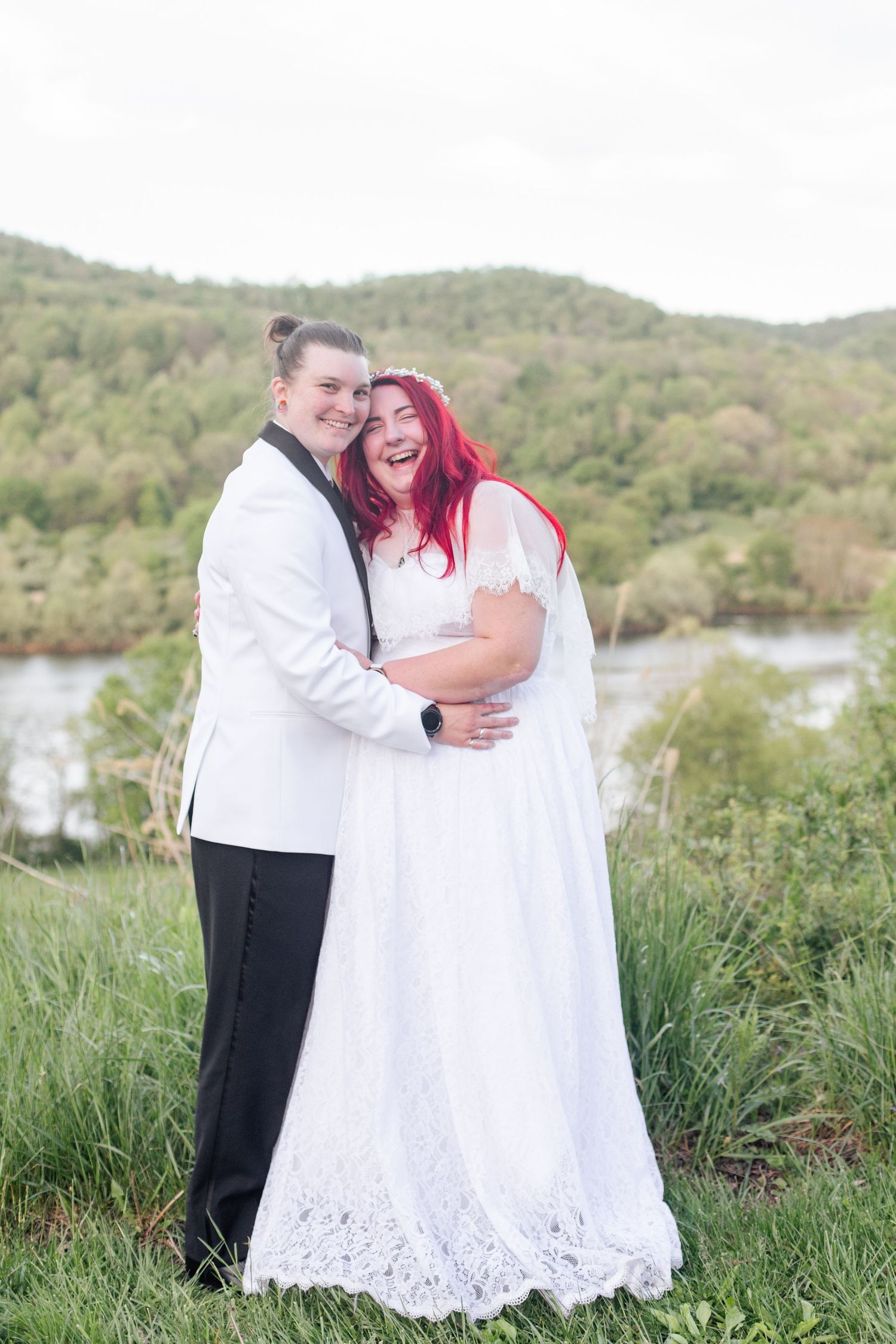 Couple embraces in grassy field with mountain lake scenery in the background.