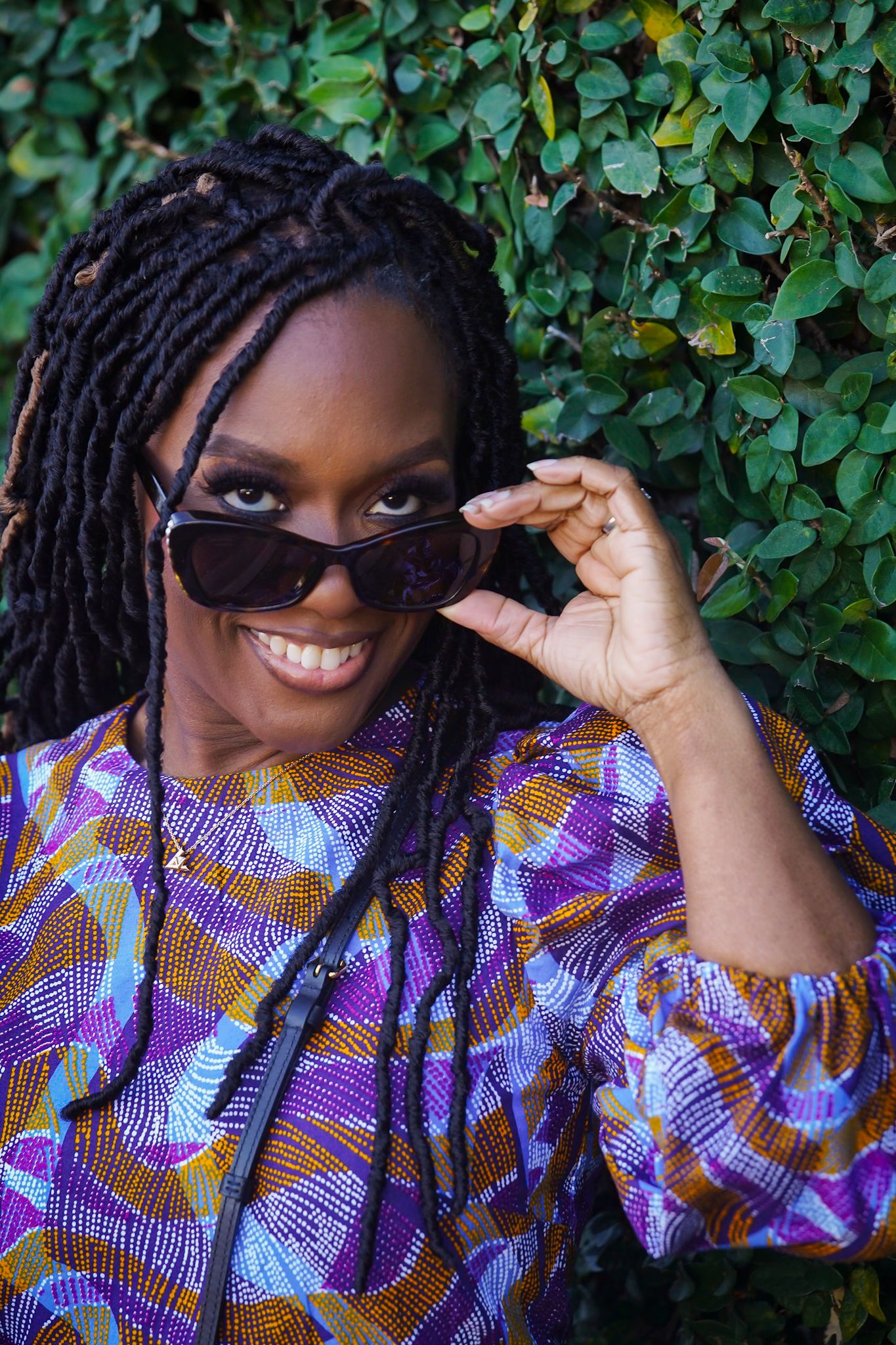 Vibrant portrait with colorful African print dress and sunglasses against green foliage.