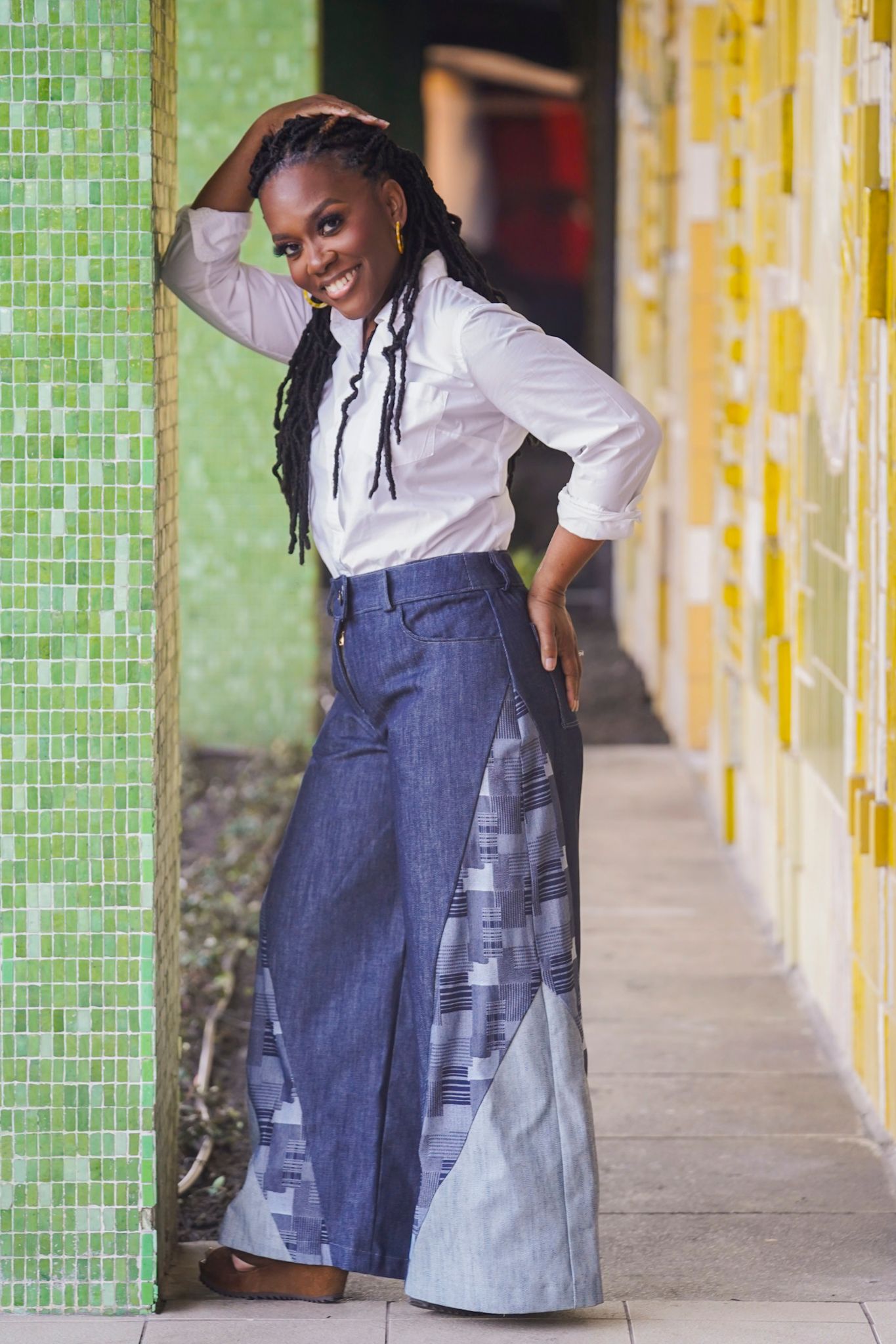 Stylish pose in wide-leg patchwork denim pants and white button-up shirt against colorful wall.