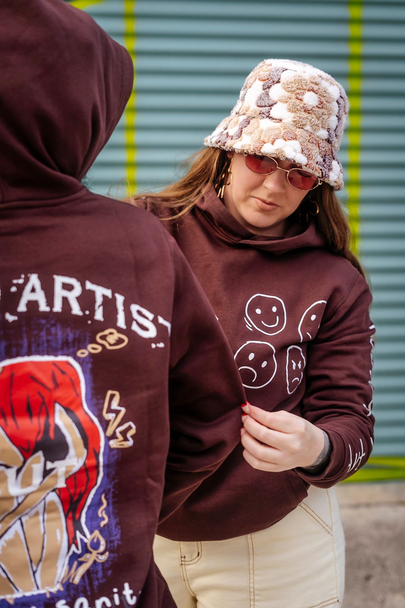Person wearing brown hoodie with smiley faces and floral bucket hat against industrial backdrop.