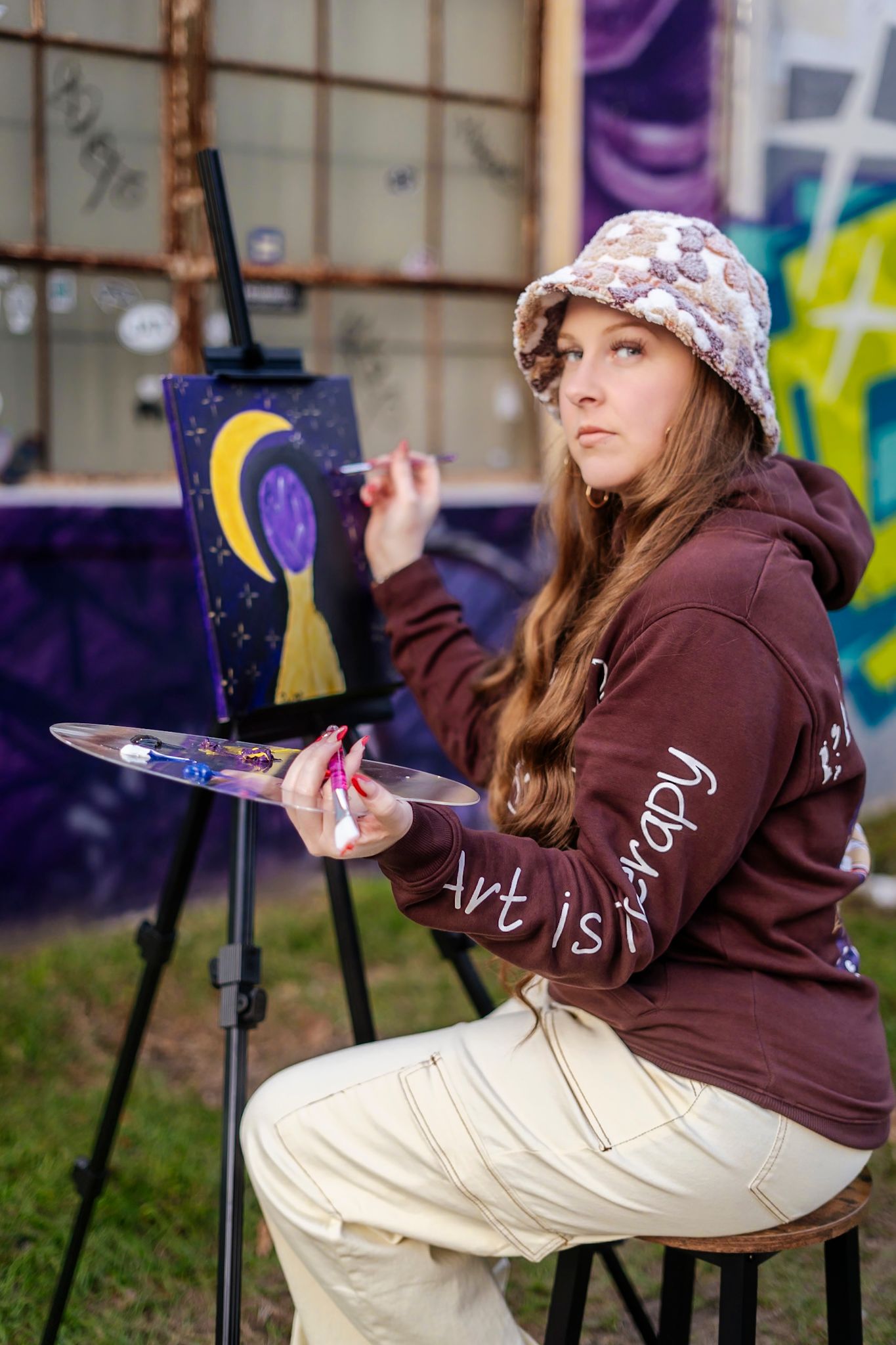 Artist in brown hoodie painting moon scene on easel outdoors.