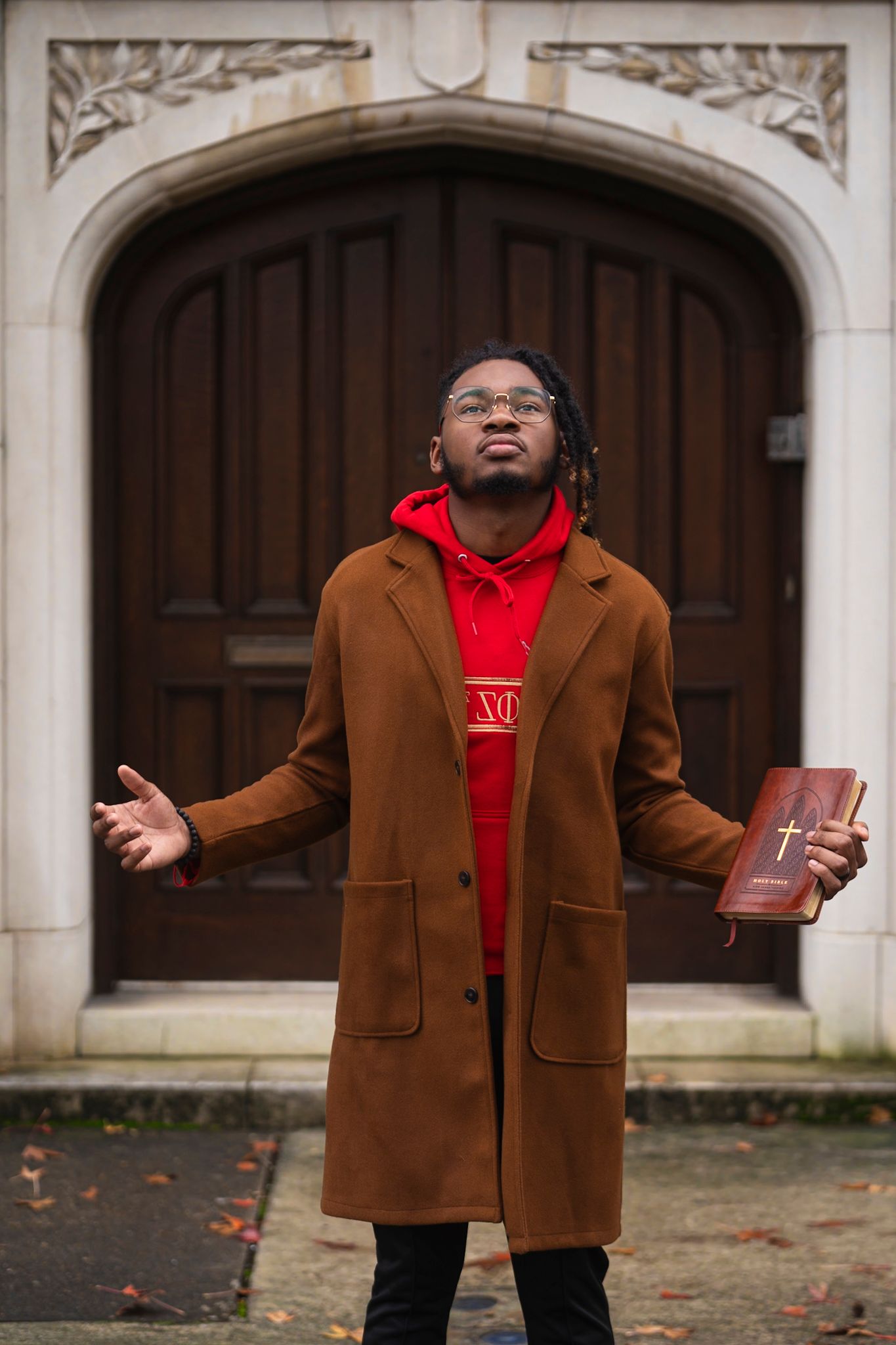 Person in brown coat and red hoodie standing in front of ornate wooden door holding a book.