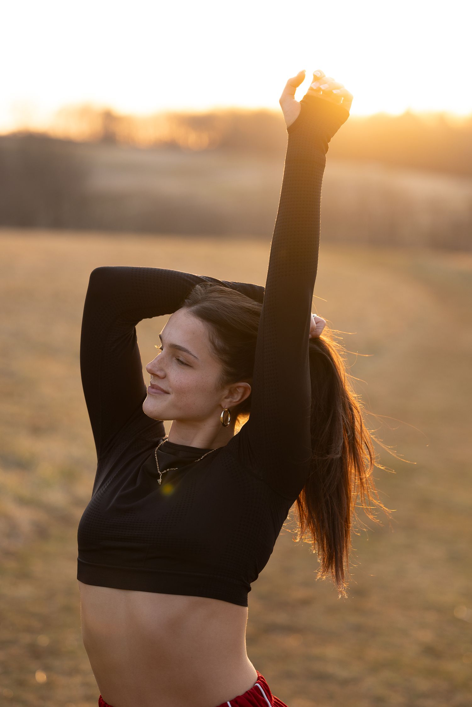 Silhouetted figure with ponytail stretches joyfully against golden sunset in black crop top outdoors.