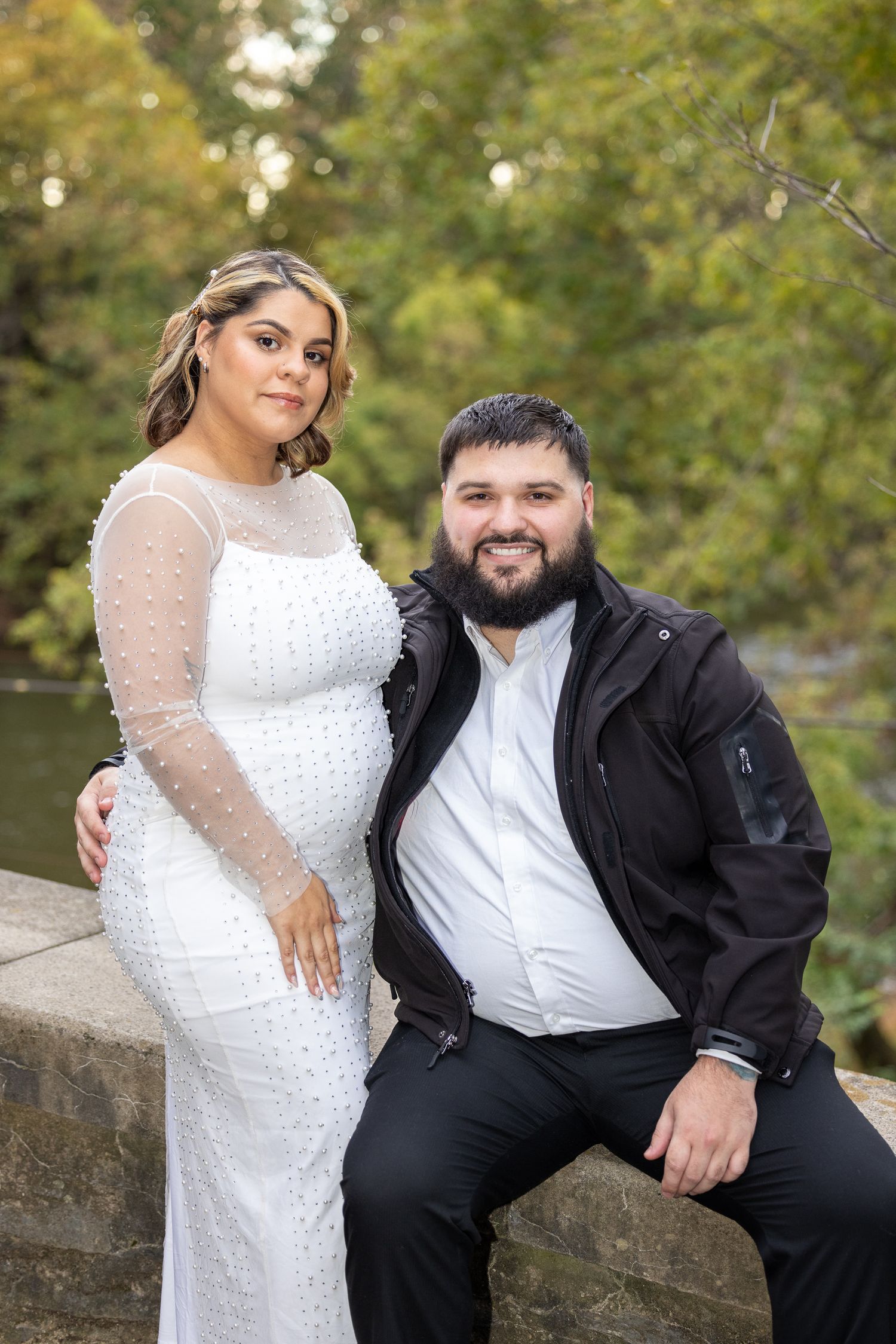 Wedding portrait of couple in white dress and black suit sitting on stone wall with autumn trees.