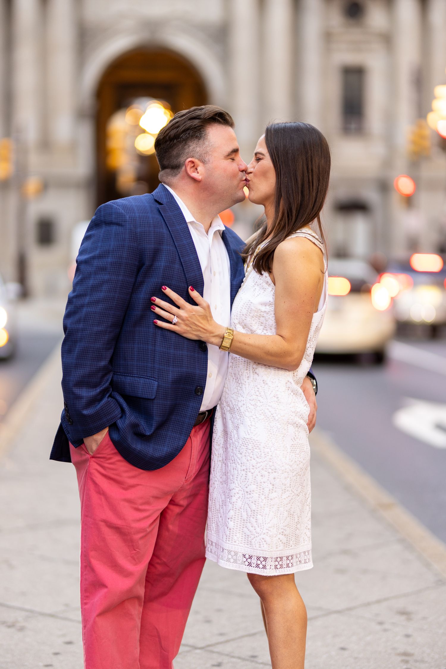 Couple kisses on city street with man in pink pants and woman in lace white dress.