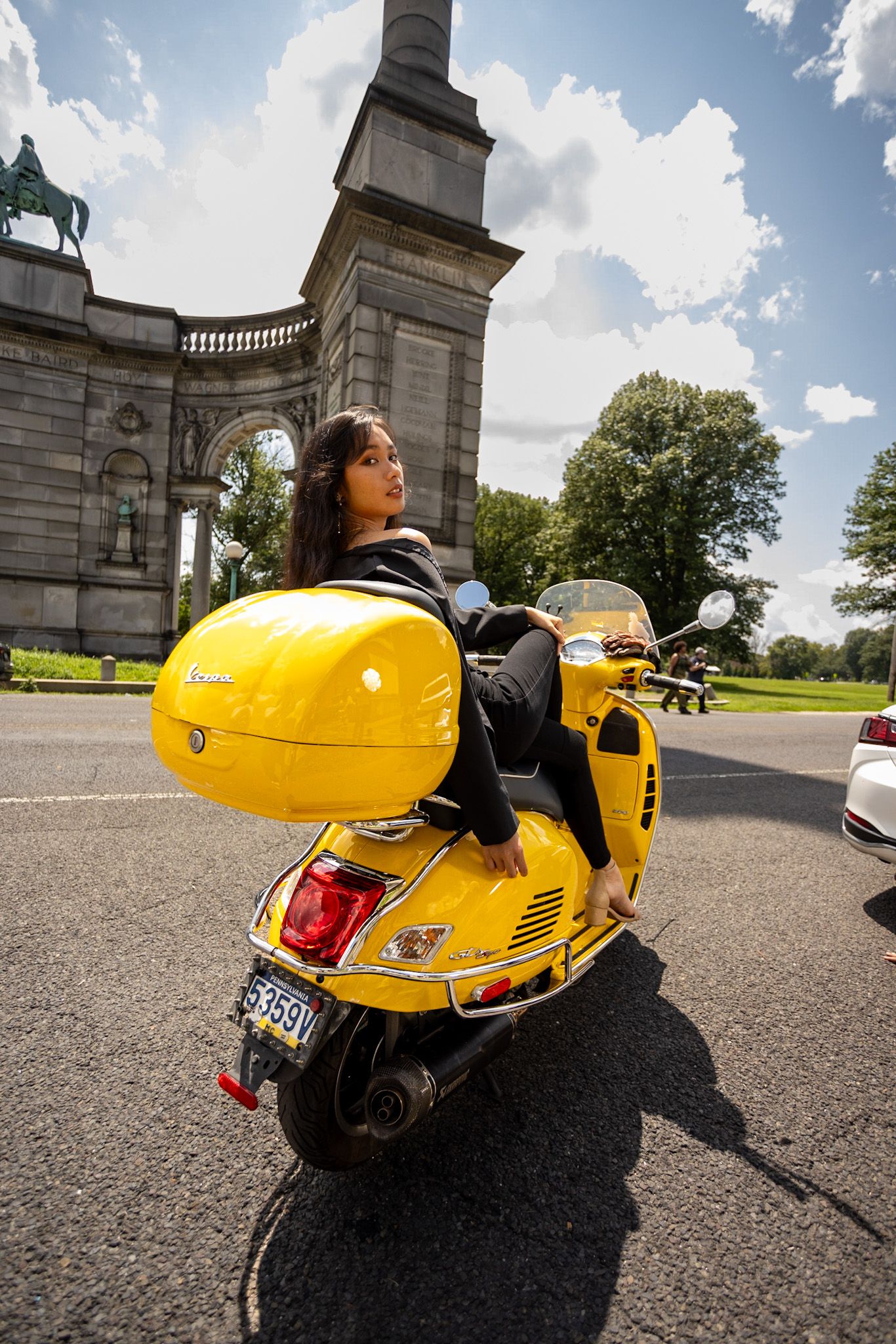 Stylish rider poses on bright yellow Vespa scooter near historic building.