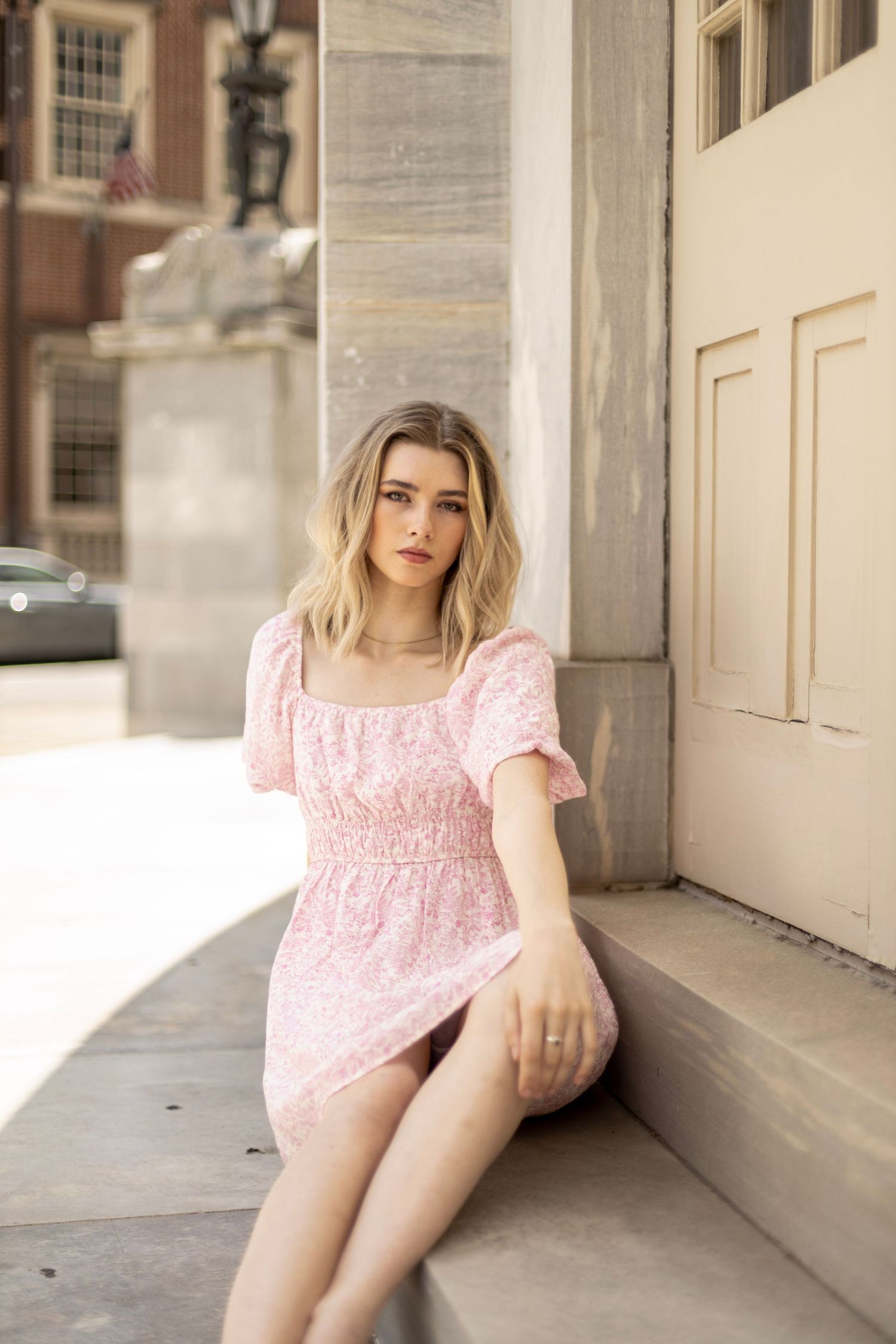 Woman in pink floral dress sits on stone steps near classical architecture.