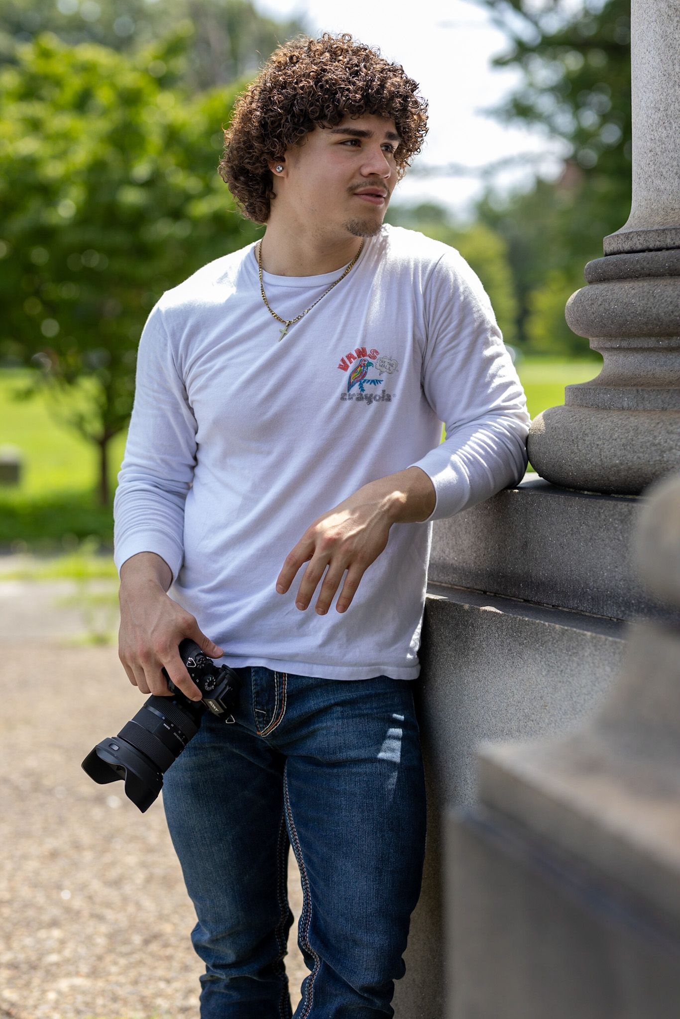 Photographer with curly hair holding a camera while wearing a white long sleeve shirt and jeans by a stone pillar.