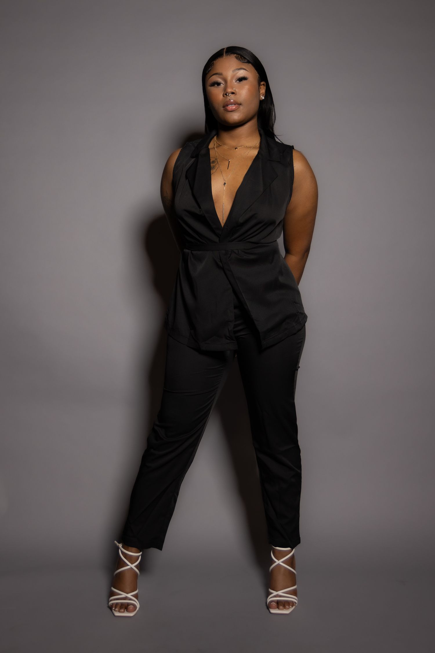 Stylish fashion portrait of person in black jumpsuit and white strappy sandals against gray background.