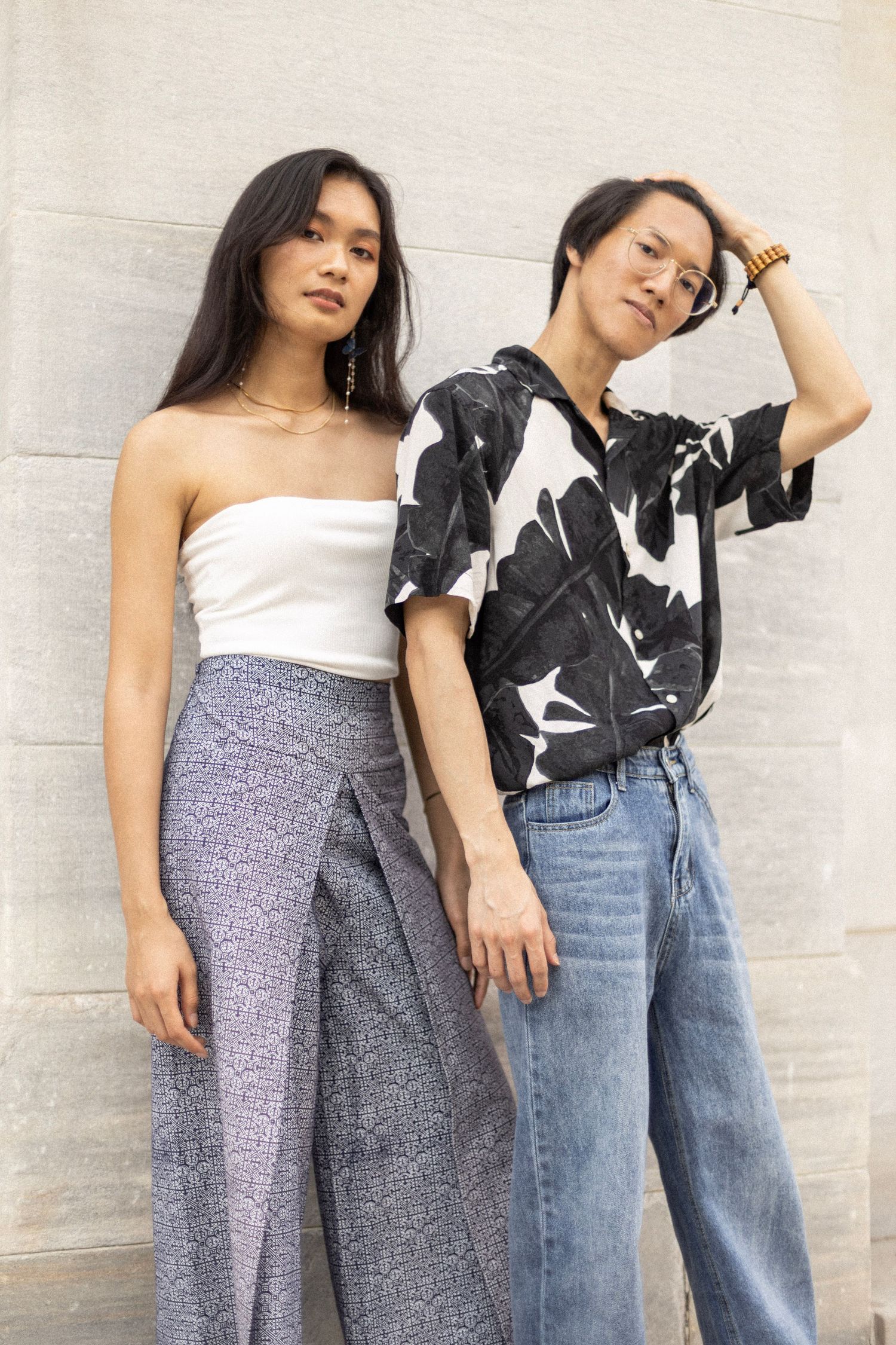 Two models pose against wall wearing contrasting outfits of white top and patterned pants.