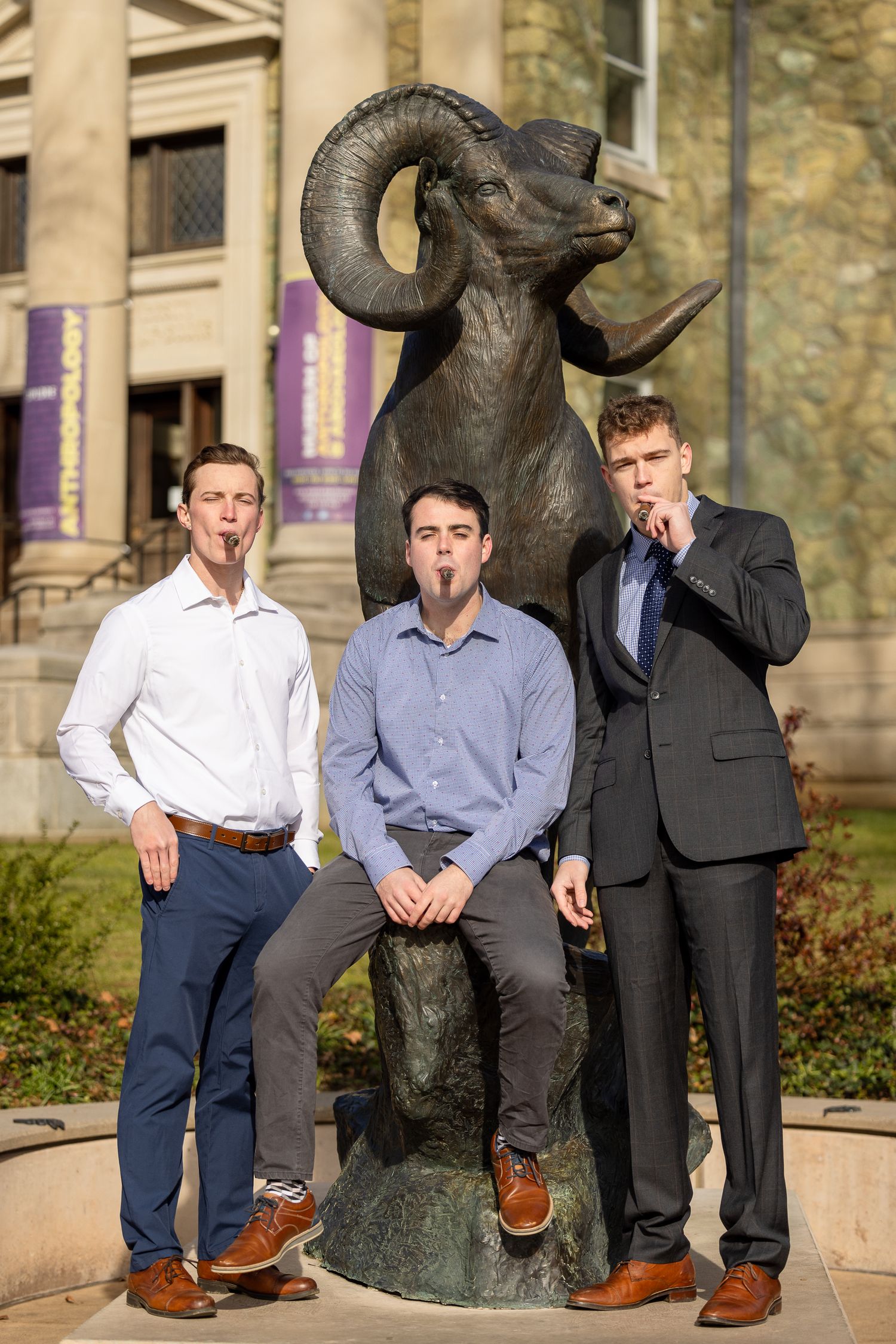 Three men pose playfully with a ram statue on a university campus.
