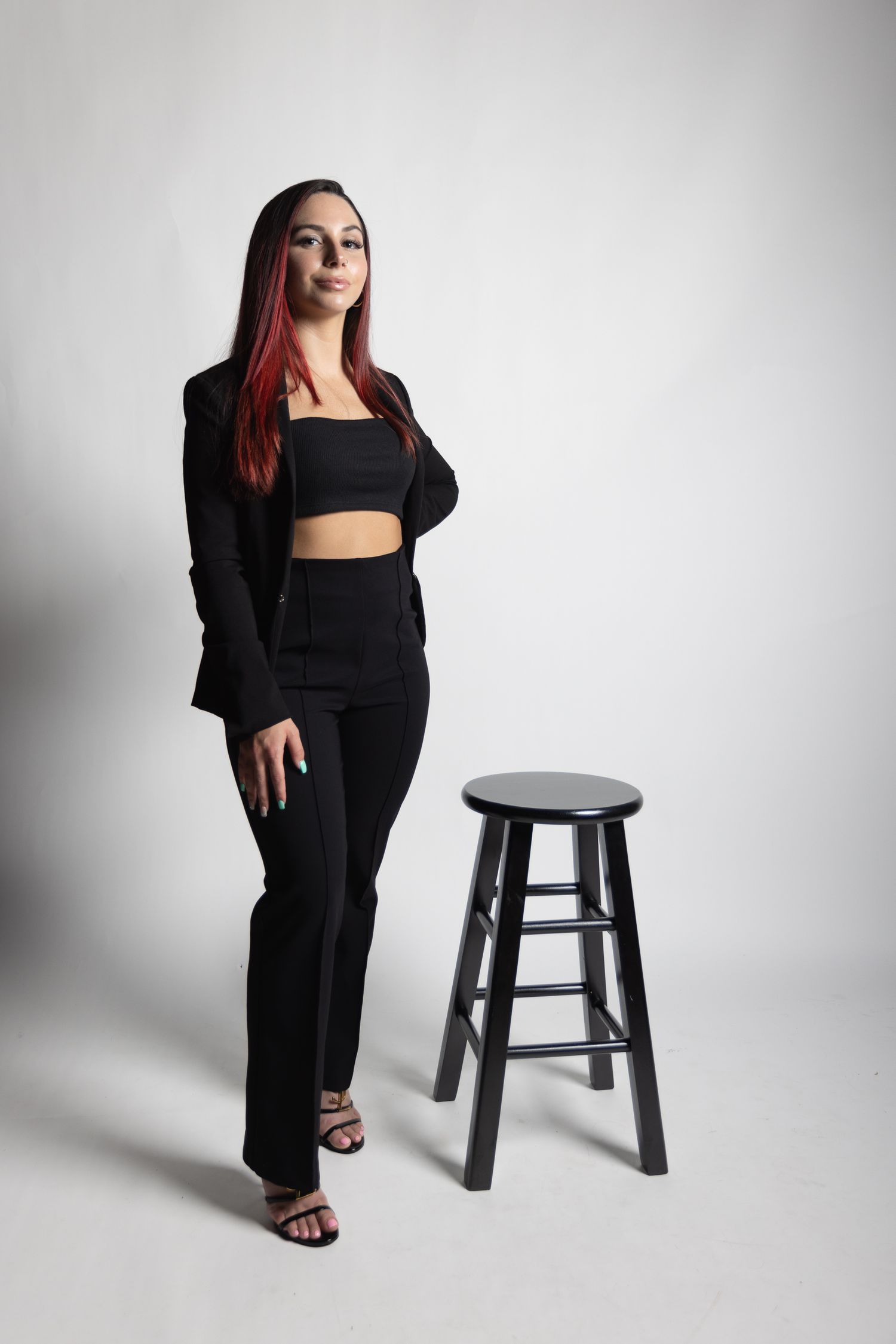 Woman in black outfit with red hair poses next to stool in studio setting.