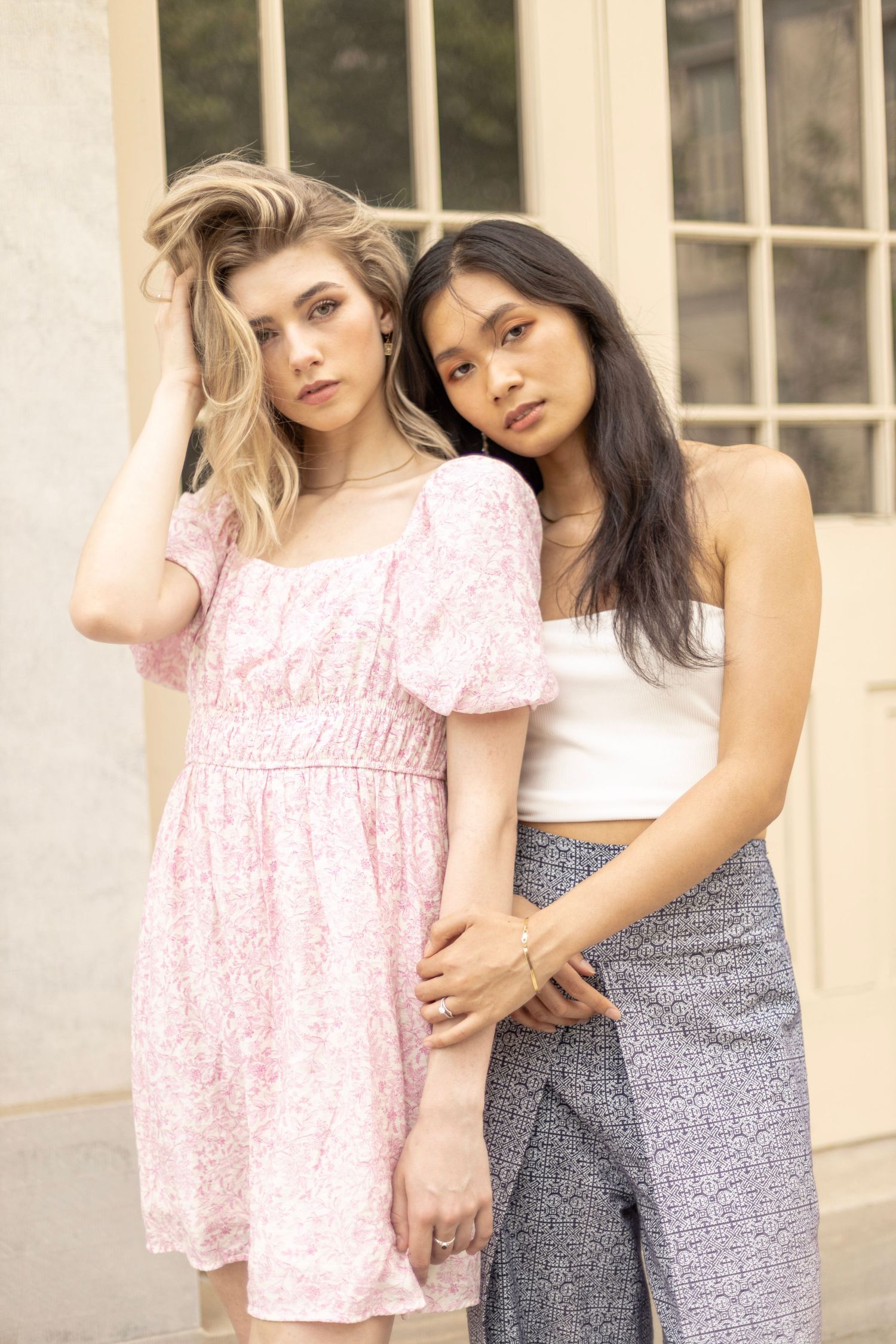 Two models pose together wearing pink dress and white top with patterned pants.