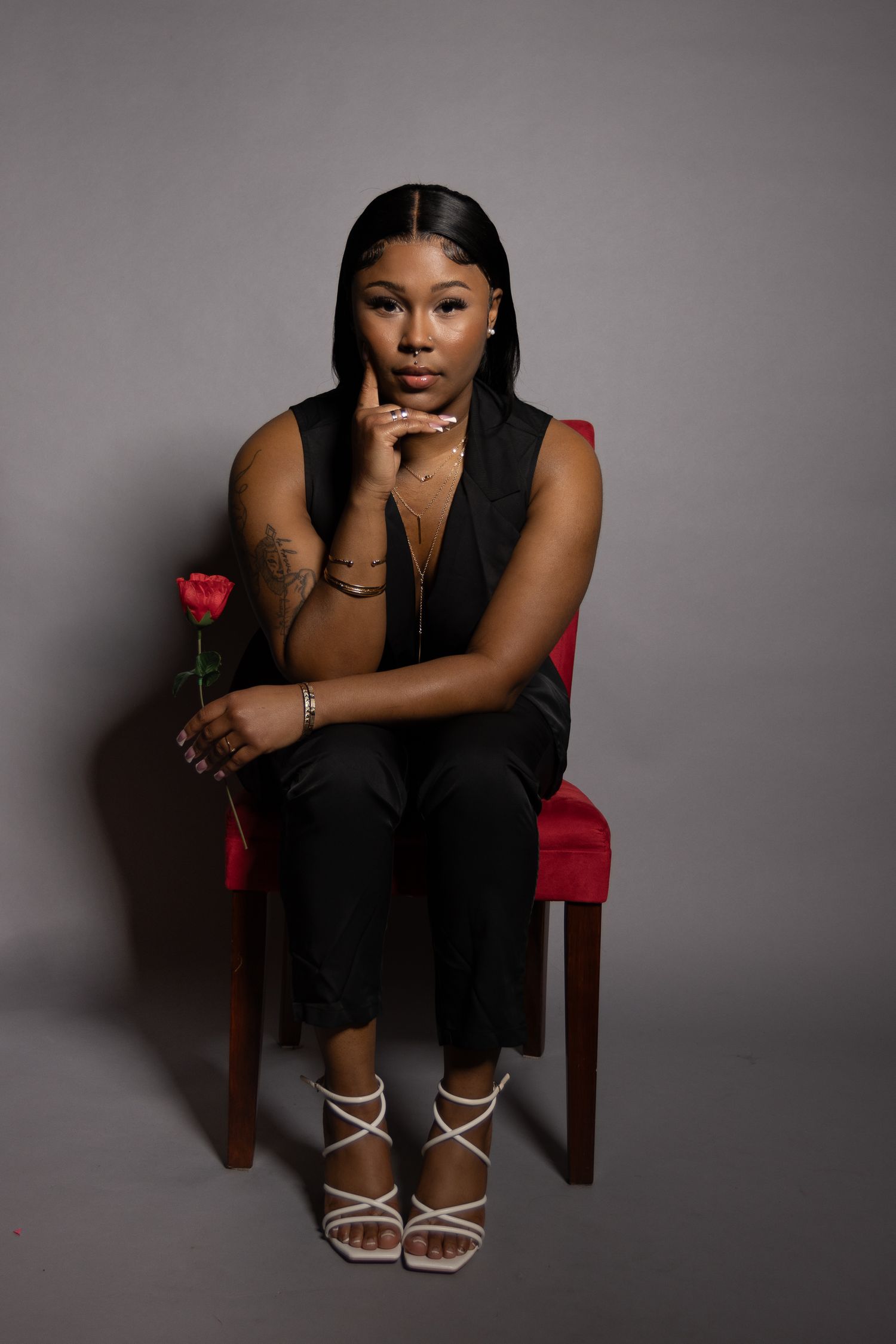Elegant portrait of person in black vest holding red rose while seated on red chair.