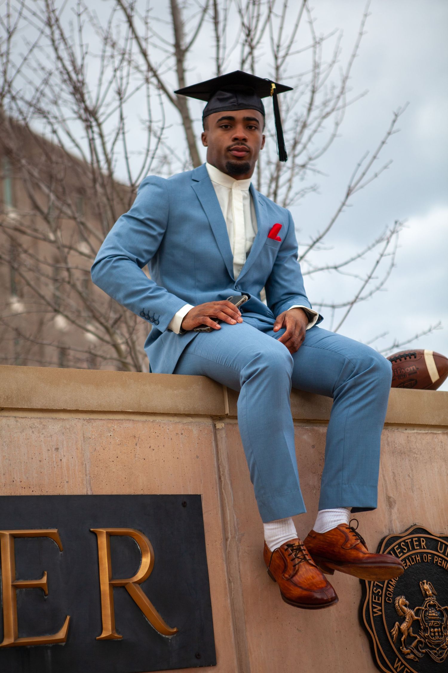 Graduate in powder blue suit sits on stone ledge wearing academic cap and brown dress shoes.