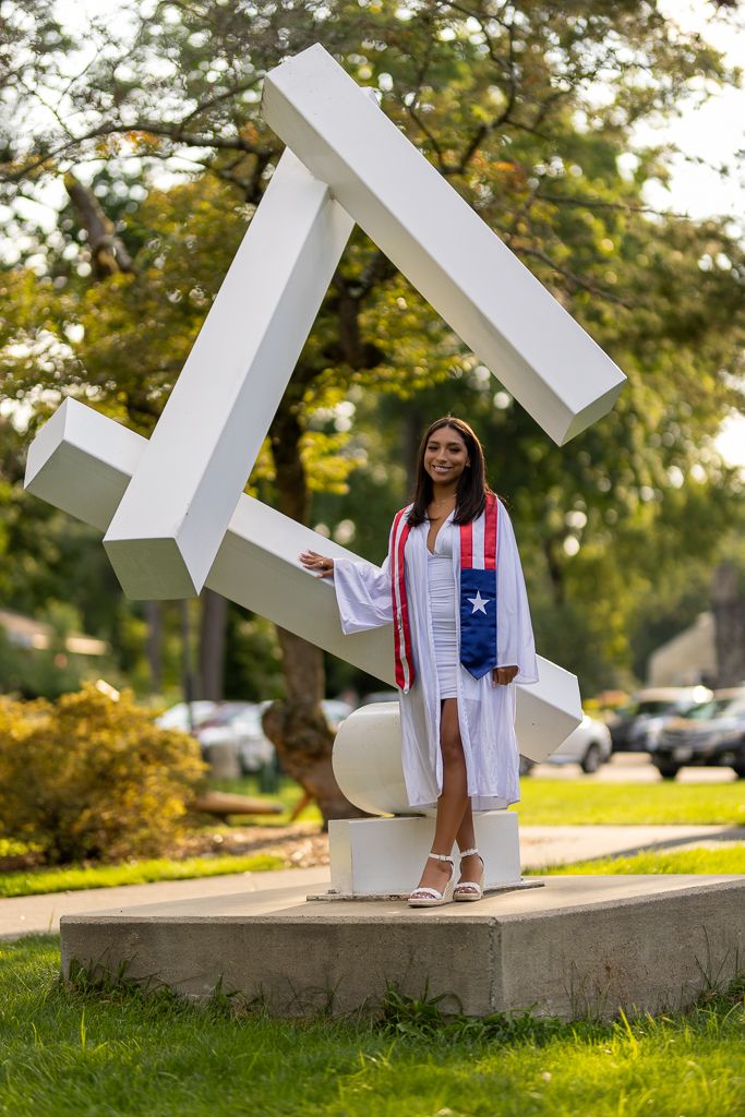 Graduate in white academic regalia poses with abstract geometric sculpture outdoors.