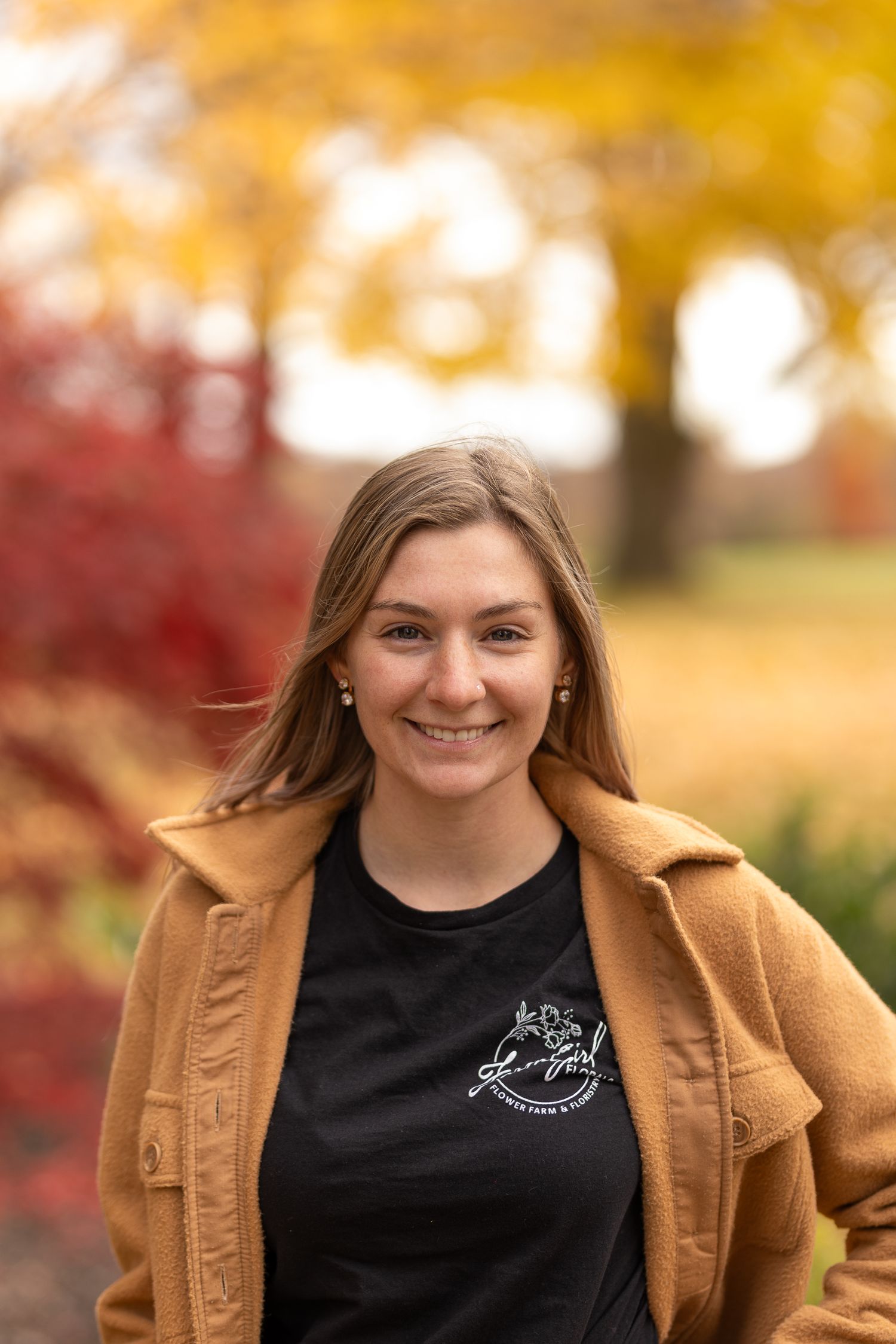 Portrait of person in tan jacket and black shirt against vibrant autumn foliage.