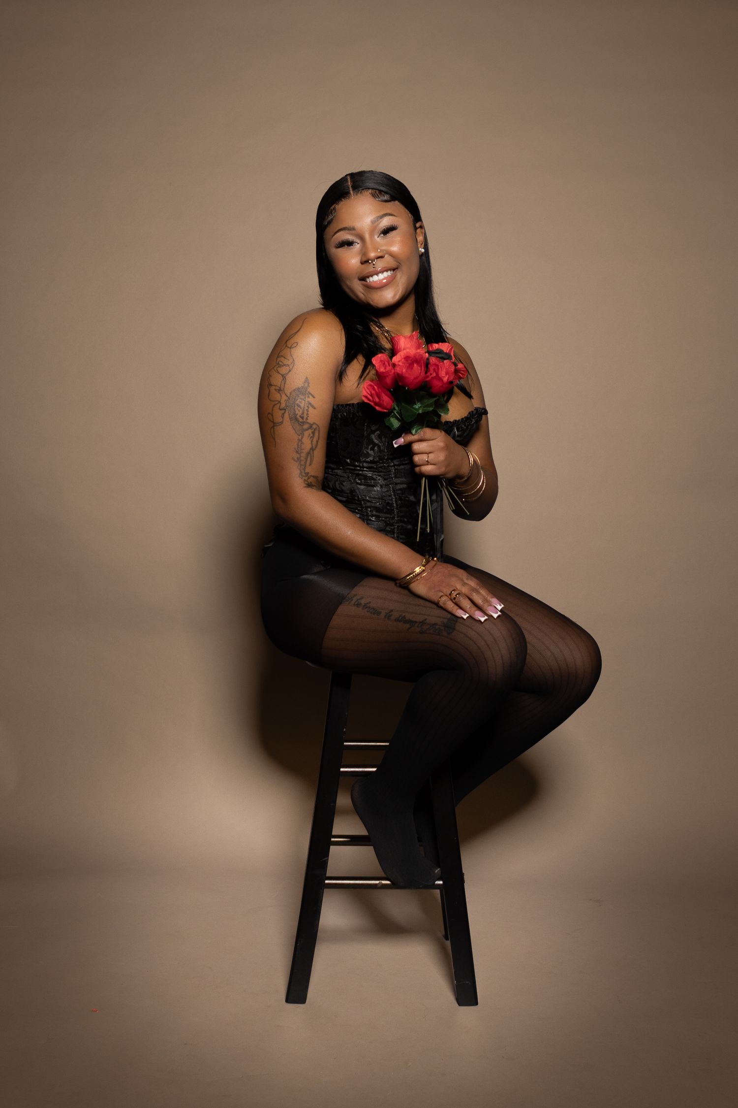 Portrait of person in black outfit holding red roses while sitting on stool against beige backdrop.