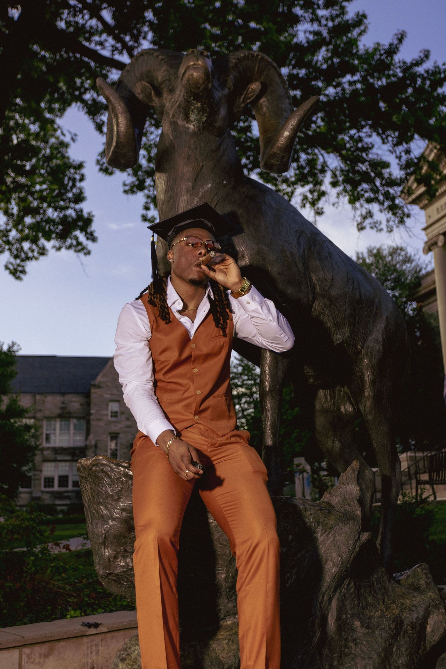 Graduate in brown vest and slacks poses stylishly in front of ram statue.
