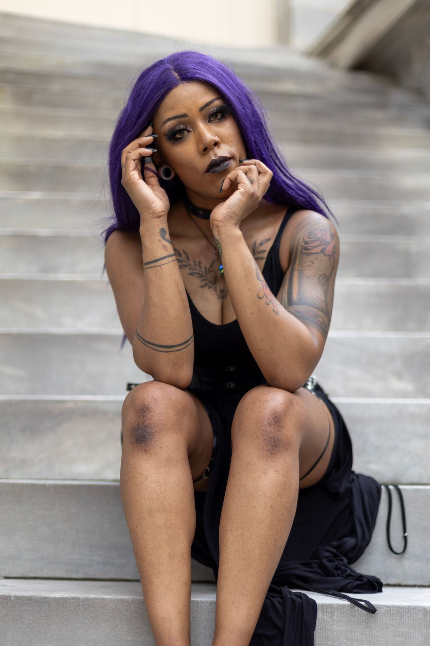 Model with purple hair and tattoos poses on concrete steps wearing black outfit.