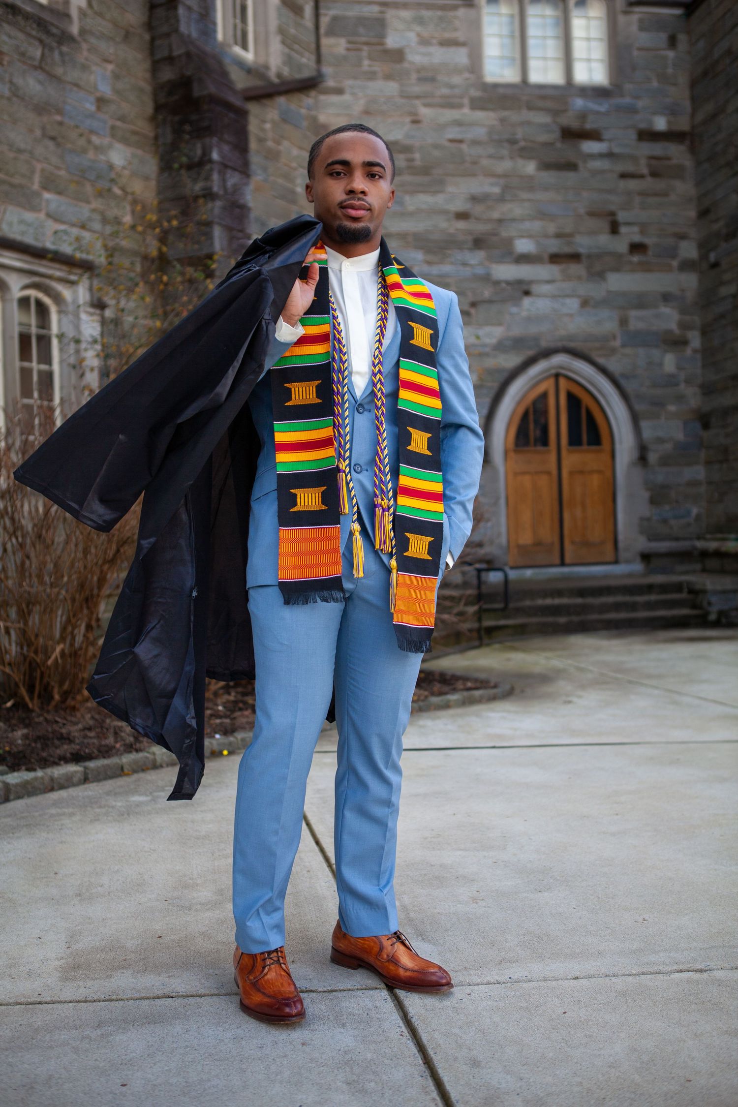 Graduate wearing traditional kente stole over light blue suit and black coat stands in front of stone building.