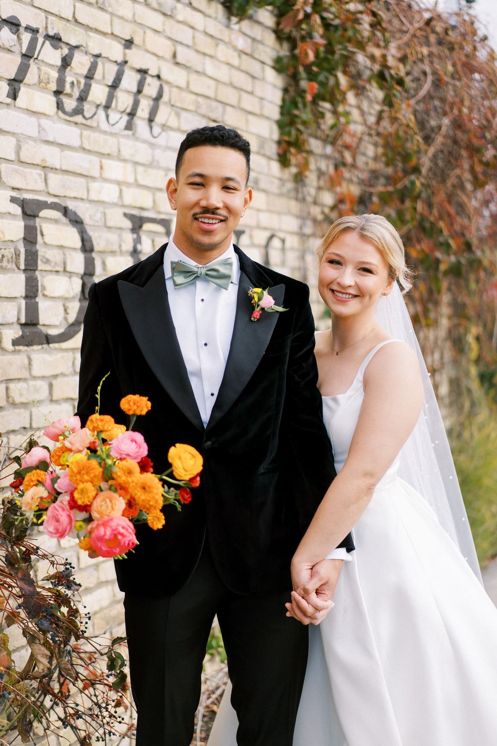 Wedding couple posing outdoors against a brick wall with vibrant orange and pink floral bouquet.