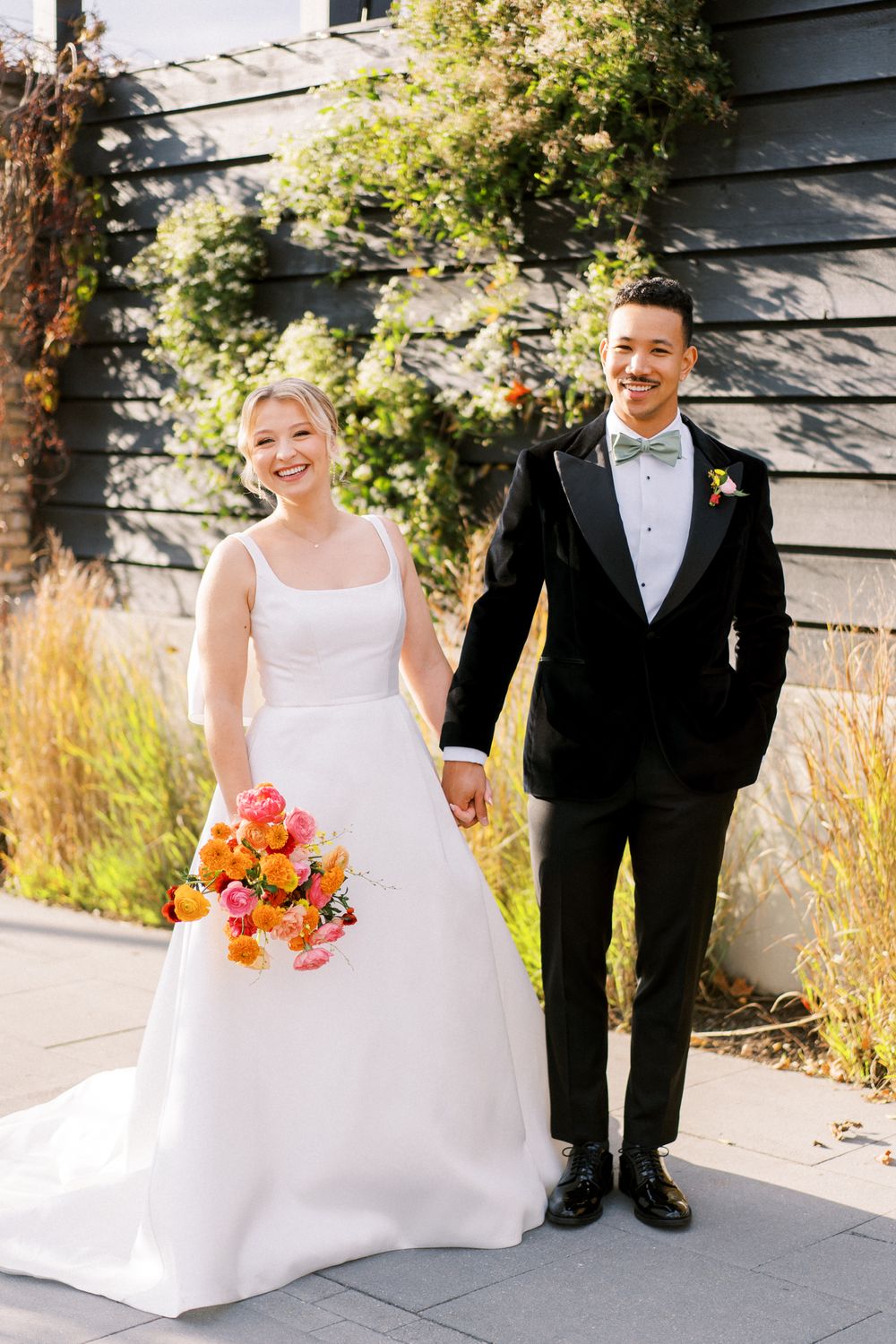 A happy couple poses outdoors in wedding attire with a bright coral bouquet against a dark building.