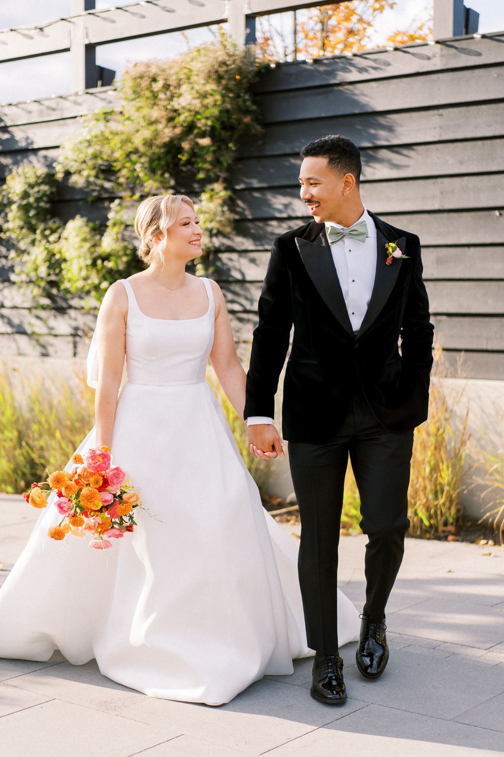Bride in classic white gown holds vibrant orange bouquet while walking with groom in black suit.
