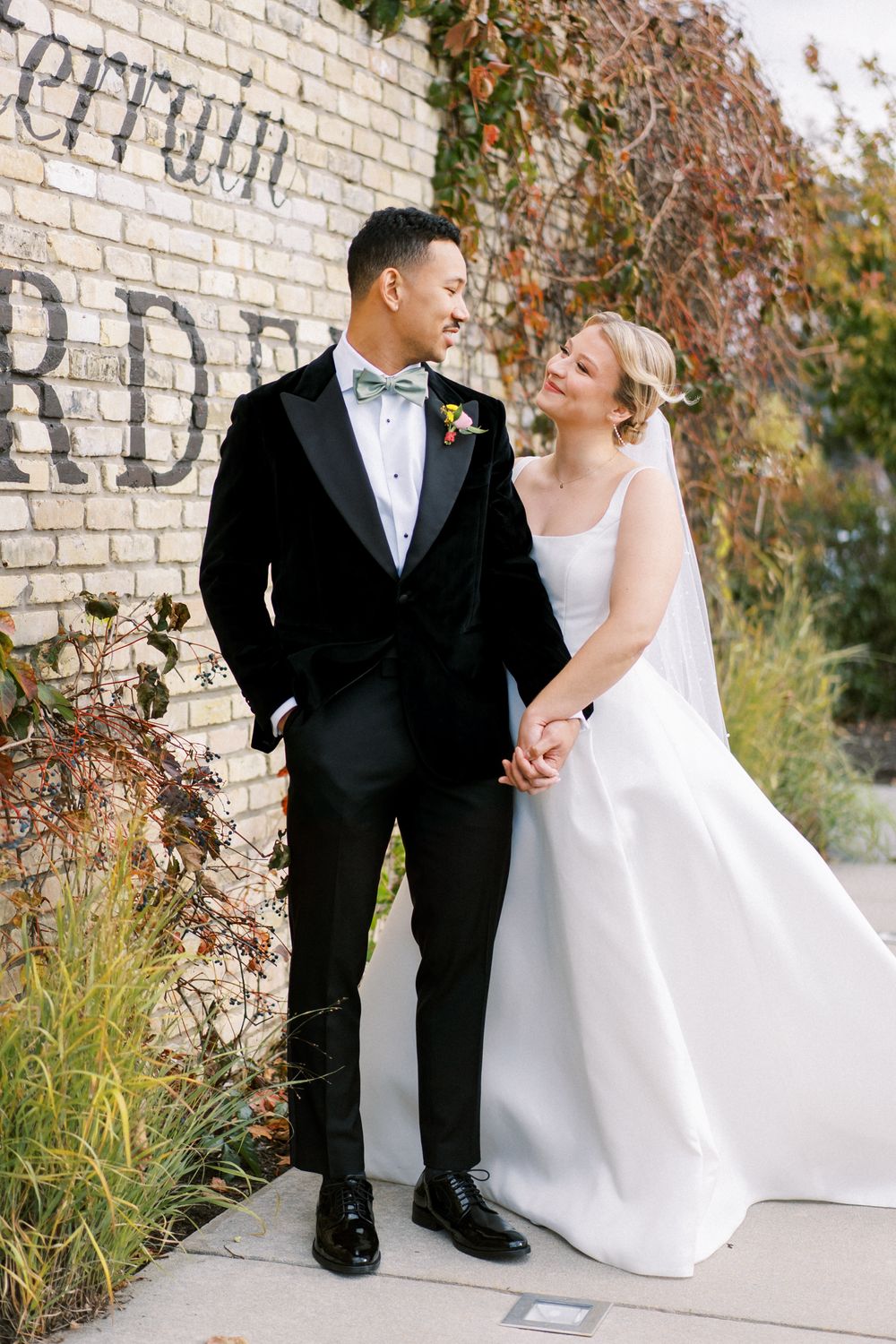 Wedding couple in elegant attire posing against brick wall with autumn foliage.