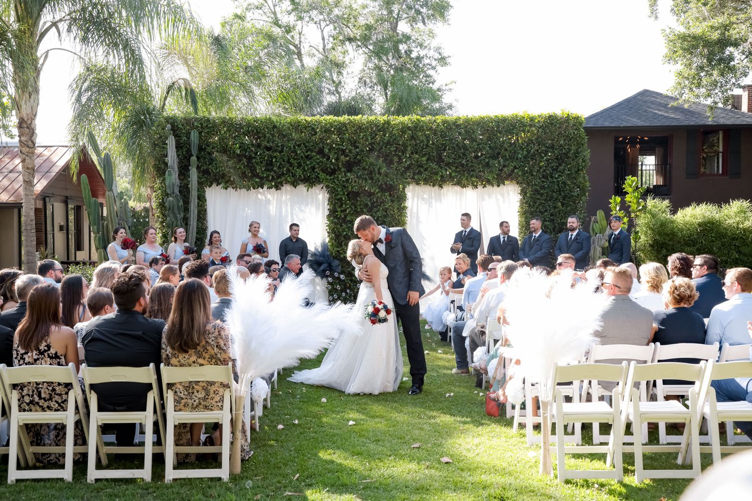 Outdoor garden wedding ceremony with white chairs and floral wall backdrop.