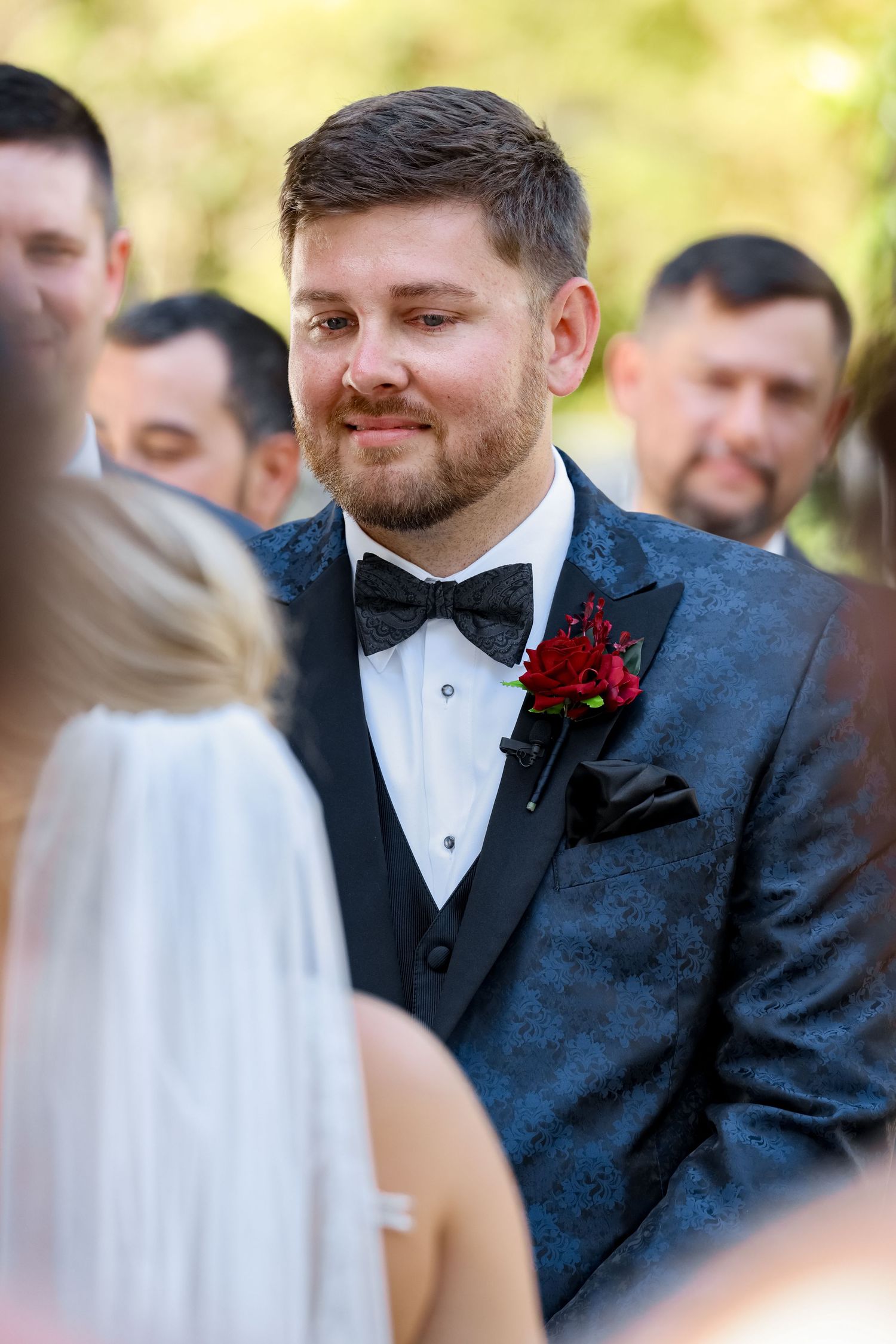 Groom in navy tuxedo with red boutonniere smiling during outdoor wedding ceremony.