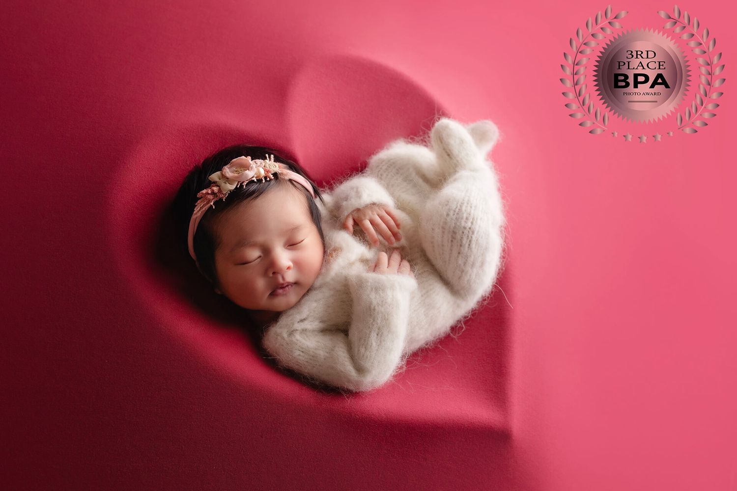 A sleeping newborn in white knit clothing rests in a heart-shaped prop against red backdrop.