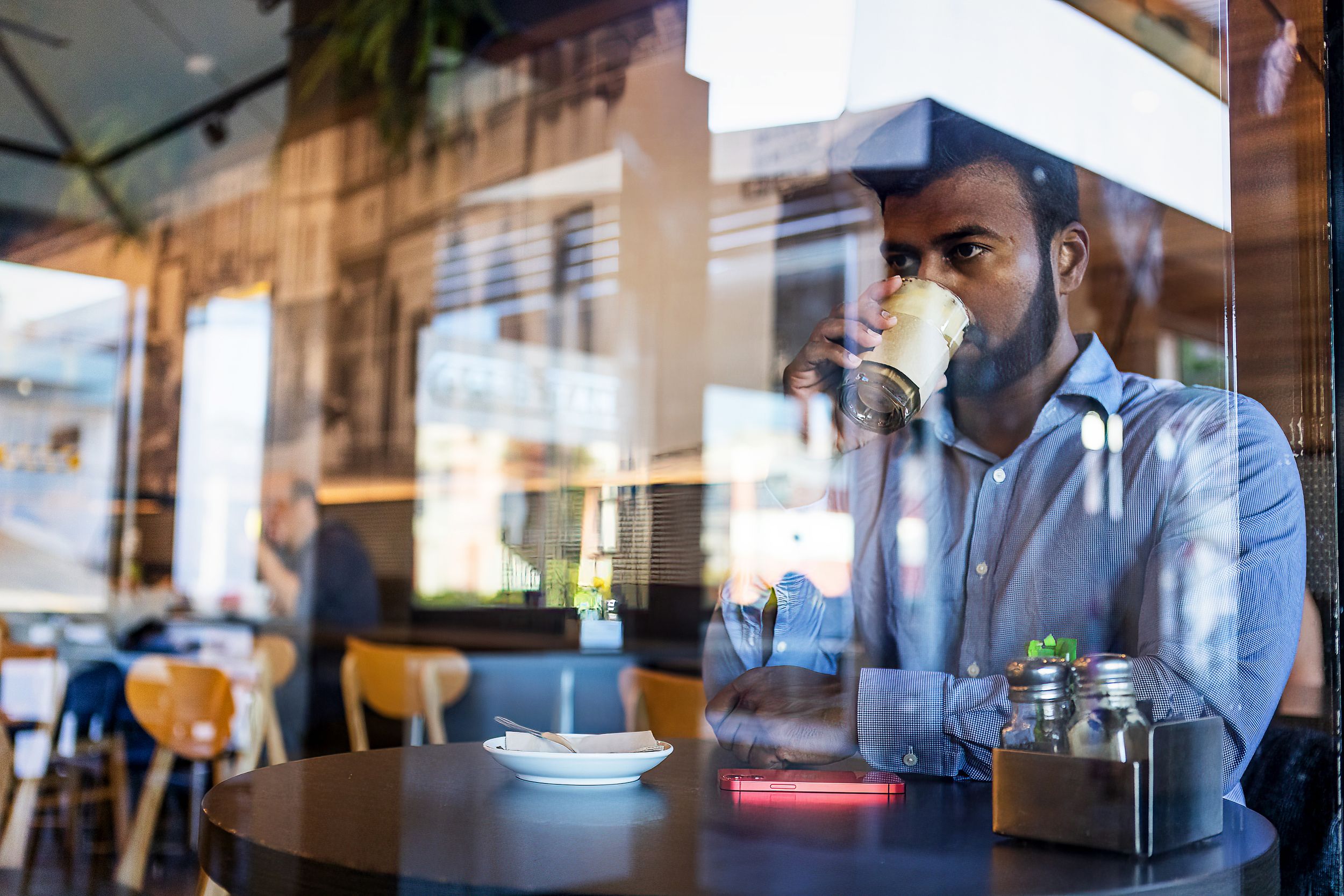 An Indian man with a beard drinks coffee in a cafe, seen through a glass storefront that reflects the street outside.